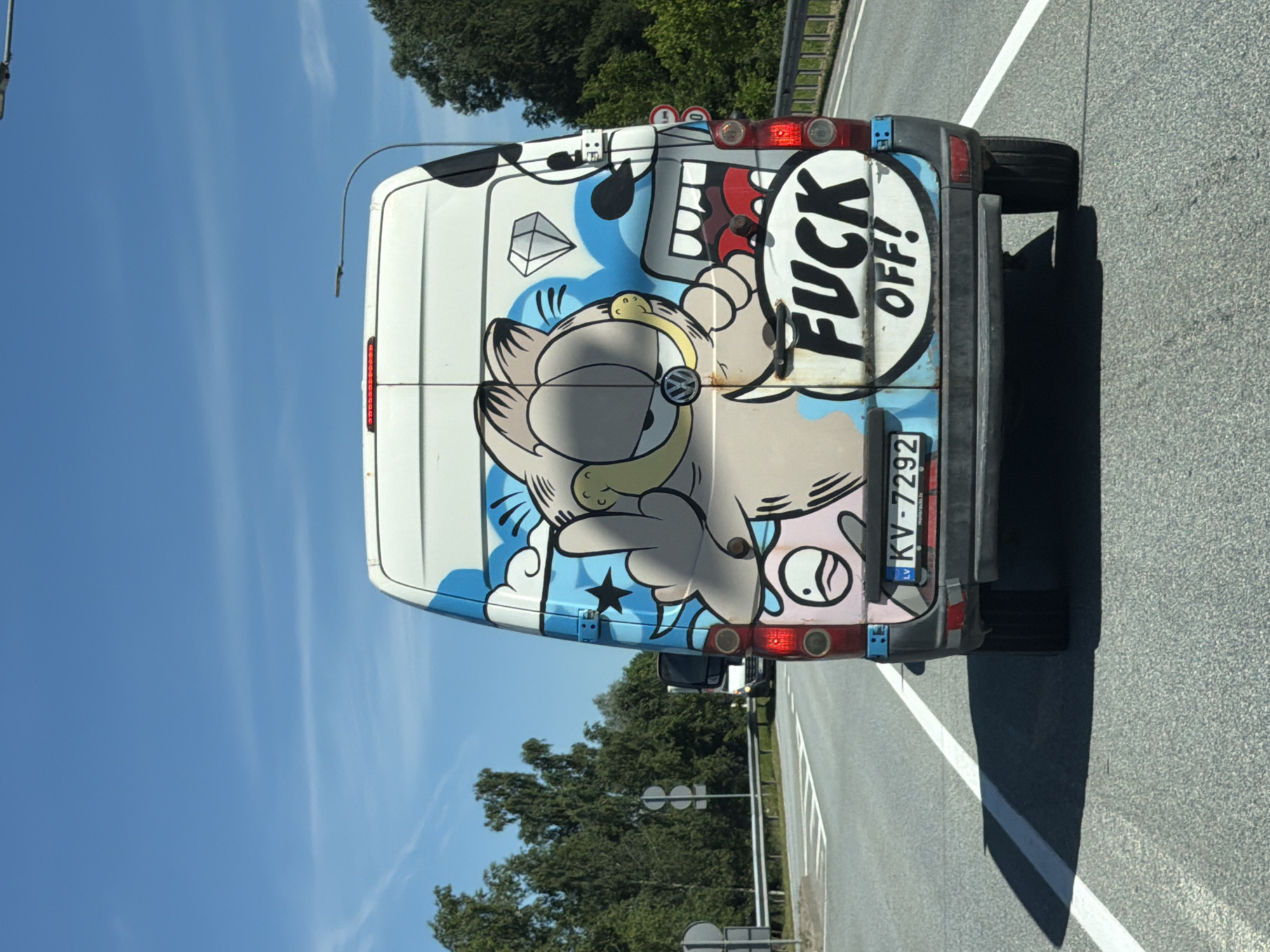 A white van with a cartoonish Garfield character painted on the back is seen from behind, driving on a paved road. The Garfield character is depicted with a middle finger raised. Below the character, the words "FUCK OFF!" are emblazoned in large black letters. The van has a license plate that reads "KV-7292 LV". The environment appears to be a sunny day with a blue sky and some clouds. Trees are visible on the side of the road. The van is in motion, and the image is taken from another vehicle, indicated by the perspective and the shadow of the camera vehicle on the road. The scene is likely in Latvia, as suggested by the license plate.