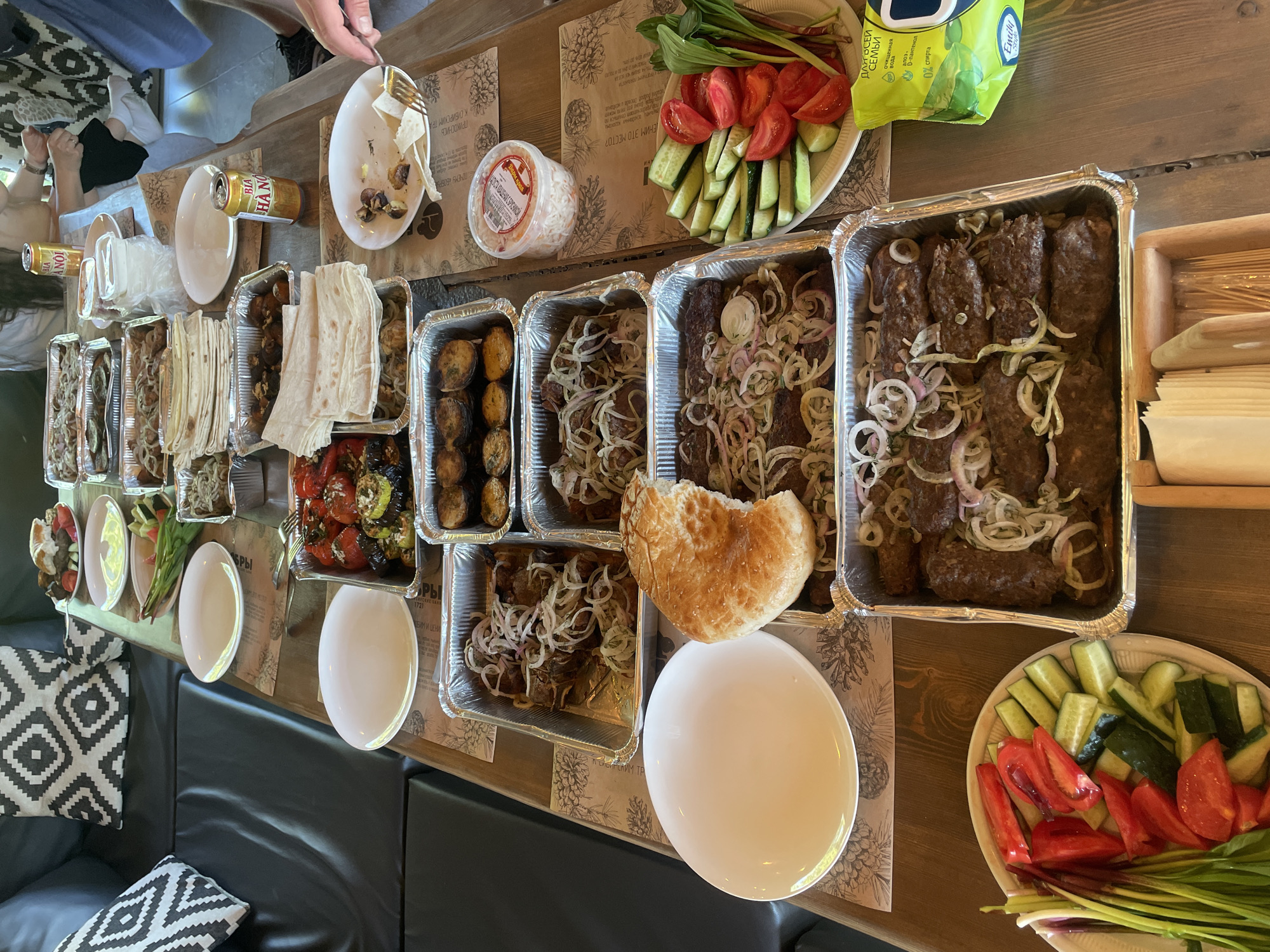 This overhead shot captures a rich meal laid out on a wooden table, suggesting a casual gathering or feast in Drokino, Russia. The table is laden with various dishes, predominantly served in rectangular aluminum foil containers, indicative of catering or takeout.
The main dishes appear to be grilled meats, possibly lula kebabs or kofte, generously topped with thinly sliced white and red onions, and some fresh dill. Several containers hold these meat preparations. Other foil containers contain roasted vegetables like peppers, zucchini, and tomatoes, as well as roasted potatoes or possibly mushrooms. Complementing the main courses are stacks of flatbread, likely lavash, and a large round piece of hearth bread, partially broken.
Fresh vegetables are arranged on white plates: sliced tomatoes, cucumbers, green onions, and red leafy greens. A clear plastic container holds what appears to be pickled or salted fresh cabbage. Beverages include several cans of "BIA HANOI" beer.
One person's hand, holding a fork, is visible in the upper left, reaching towards a partially eaten plate with some food remnants and flatbread pieces, suggesting the meal is underway. Another arm is visible further down the table.
The table itself is adorned with paper placemats featuring pinecone designs and Cyrillic text. Visible phrases include "ДЛЯ ВСЕЙ СЕМЬИ" (For the whole family) and details about a product like "0% спирта" (0% alcohol), "очищенная вода" (purified water) and "D-пантенол" from "Эндла" (Endla) on a green bag. Other placemat text includes "ПРИКОСНИСЬ К СИБИРСКИМ ТРОПАМ" (Touch the Siberian trails) and words like "БРЫ", "ОРЫ", "1721", possibly referring to "Бобры" (Beavers), which is common in Siberia. The setting includes black upholstered seating with geometric patterned pillows, providing a comfortable, modern-rustic ambiance. The natural lighting suggests it is daytime.
