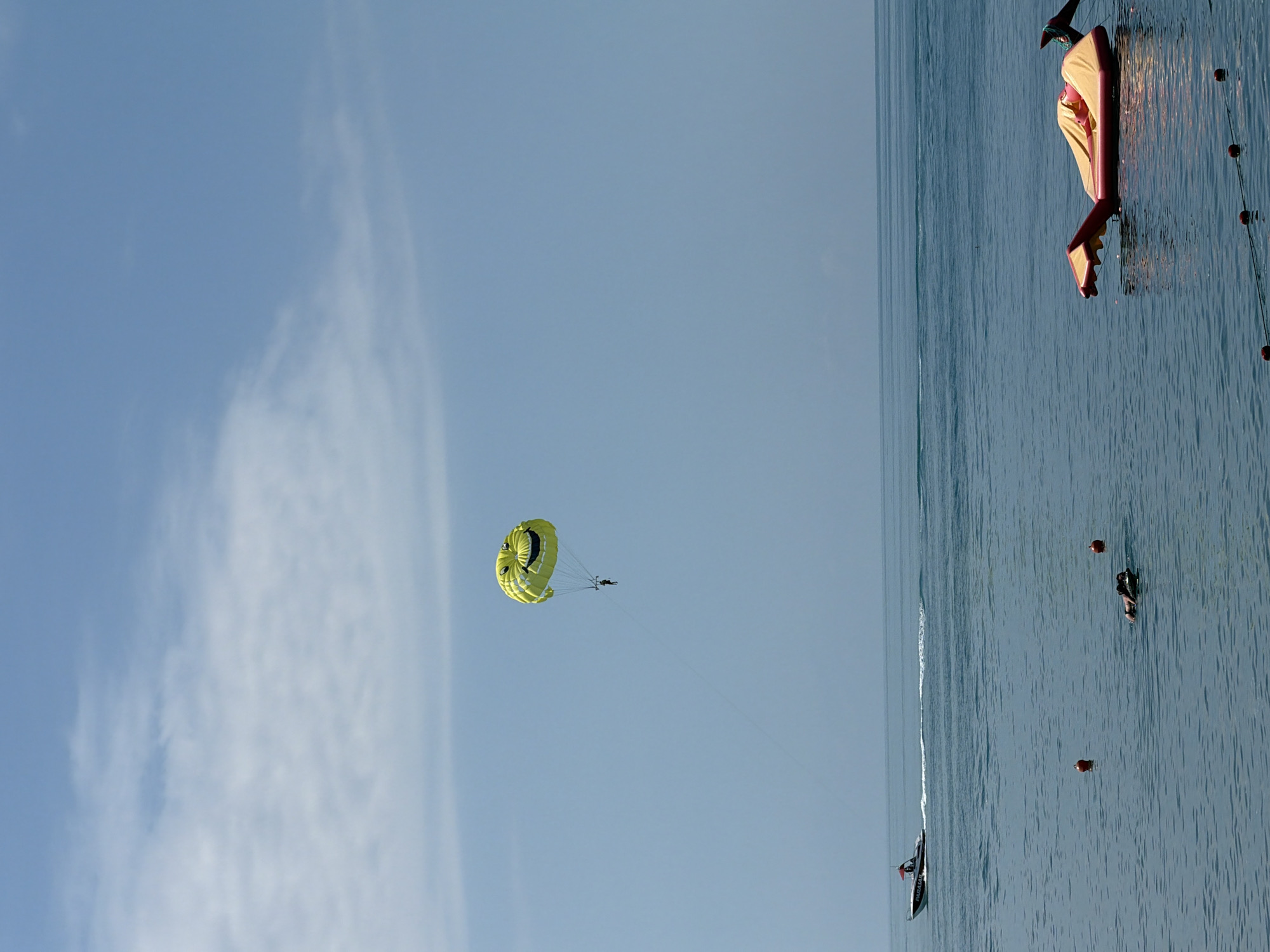 In the Black Sea off the coast of Sochi, Russia, a person is parasailing under a bright yellow parachute with a smiley face design. The parachute is high in the sky, with a thin tow rope connecting it to a boat visible in the distance. The boat is moving at a good speed, creating a wake. In the foreground, an inflatable watercraft, shaped like a dragon or sea creature, floats on the surface of the water. There are a few buoys scattered in the water, and a person can be seen swimming or floating in the sea. The sky is a clear blue with a few wispy clouds. The water is calm, reflecting the blue of the sky. The scene depicts a typical day of water sports and recreation at a seaside resort.