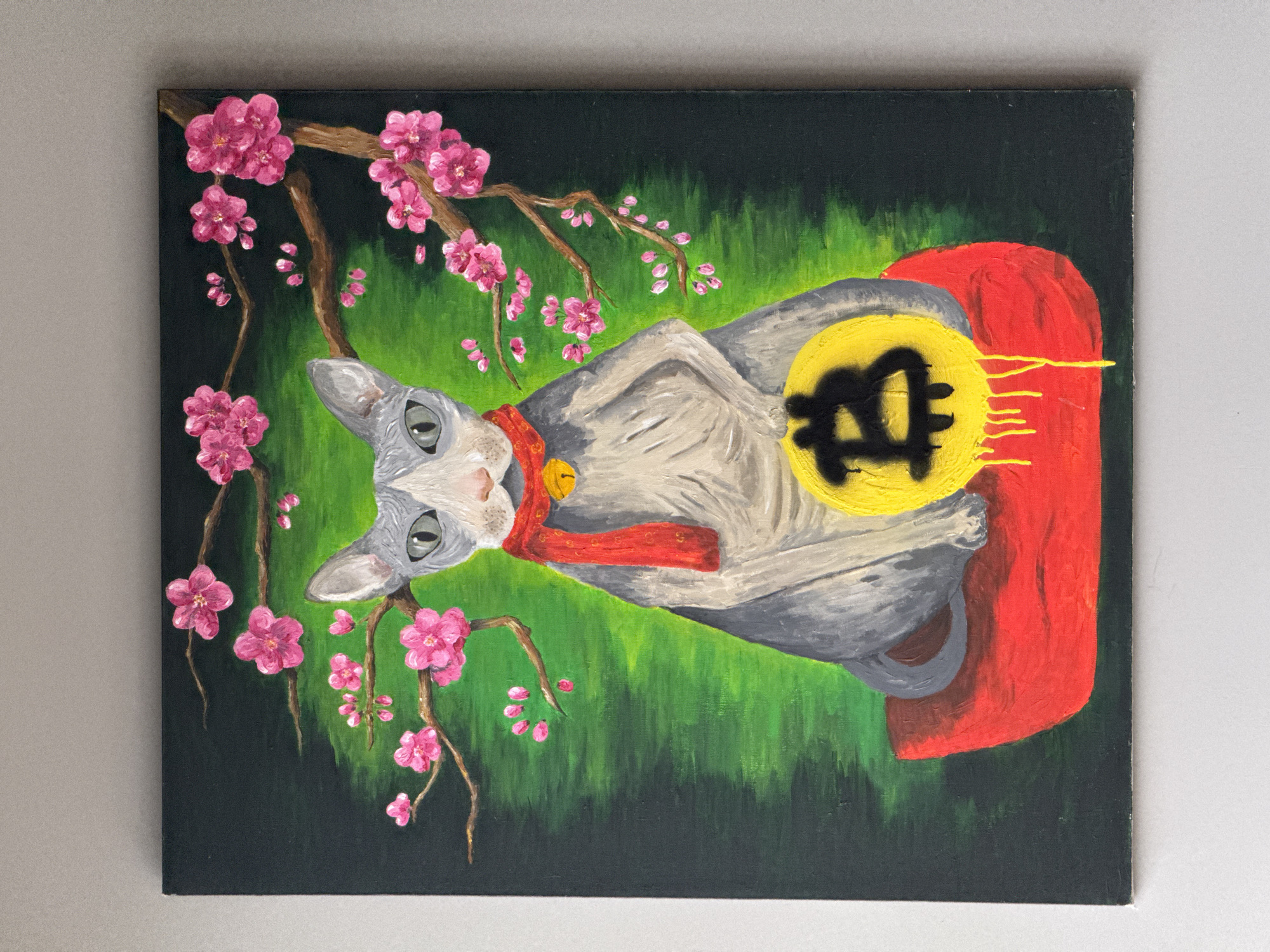 The image is a painting of a cat wearing a red cape and a red collar with a gold bell. The cat is lying in front of a backdrop of green grass with a flowering cherry blossom branch in the upper left corner. The cat has a yellow circle with a black Bitcoin symbol on its chest. The background is dark green, and the painting appears to be resting against a light grey wall. There is no information in the image to indicate a location.