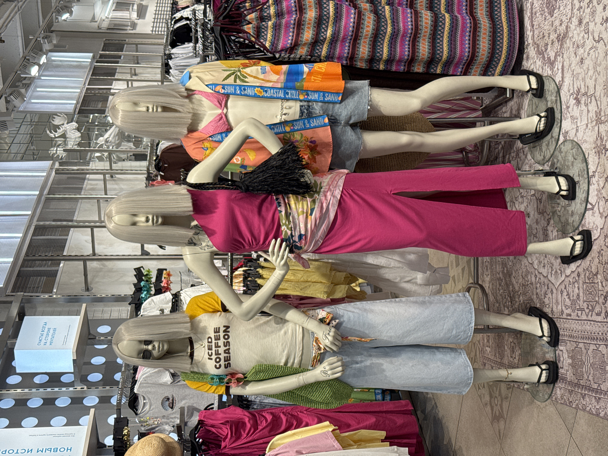 The image shows a retail store interior, likely a clothing shop, with several mannequins displaying outfits. The mannequins have platinum blonde wigs and are positioned on glass stands on a patterned floor.
The top mannequin wears a sun-themed shirt with "SUN & SAND COASTAL CHILL" printed on the band, paired with denim shorts. The middle mannequin is dressed in a pink top and matching pants, cinched with a patterned sash, and carries a black bag. The bottom mannequin wears a yellow and white "ICED COFFEE SEASON" t-shirt and light-wash denim pants.
Clothing racks filled with assorted items of apparel are visible in the background, along with store fixtures such as display boards and shelving units. The display boards feature text in Russian, including phrases like "СЧАСТЬЕ ВСЕГДА НА СТОРОНЕ МЕЧТАТЕЛЕЙ" ("Happiness is always on the side of dreamers") and "новым истор" ("new stories").
Given the Russian text, this scene is likely to be located in Saint Petersburg, Russia.