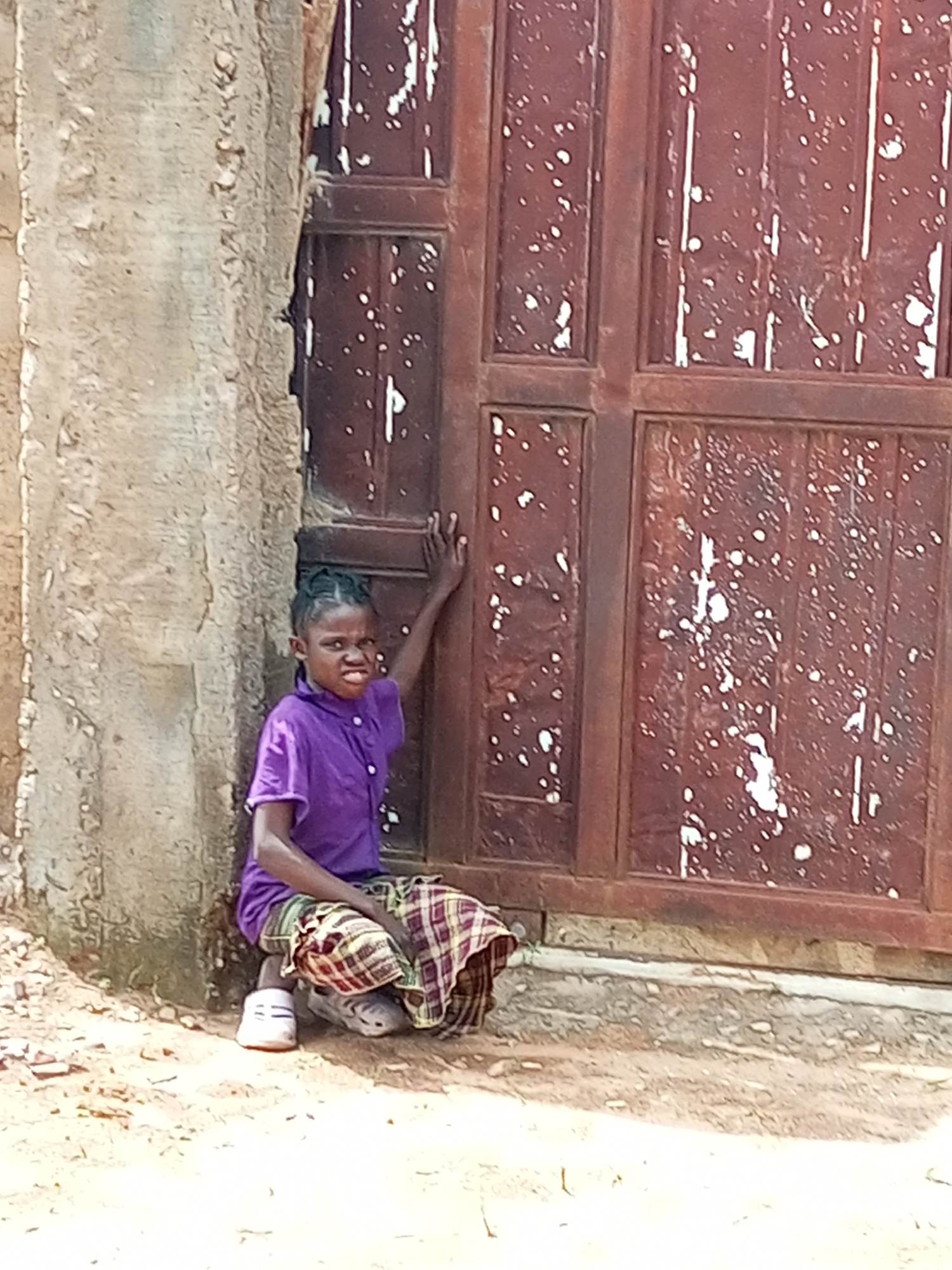 A young Black girl with braided hair, wearing a purple collared shirt and a plaid skirt, sits on the ground in front of a weathered, dark red wooden door. She is leaning against a rough, concrete pillar to her left, with her right arm extended upwards to touch the door. Her facial expression is a grimace, with her teeth bared. Her feet are clad in white sneakers. The ground around her is dirt with small stones and debris. The door, which appears to be a large double door, has a distressed finish with white paint splatters. The scene is outdoors, likely in an urban or semi-urban environment given the concrete pillar and building structure. The lighting suggests it is daytime. There are no other people or animals visible in the image.