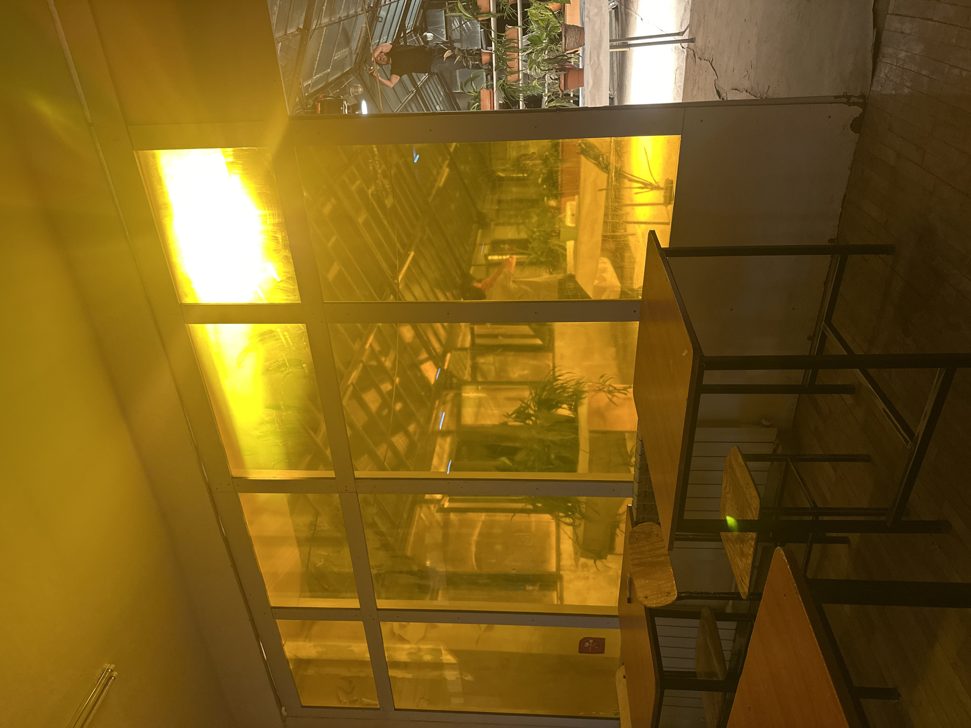 The image depicts an interior space, likely a café or restaurant, in Yerevan, Armenia. A section of the room features a large window wall with a glass roof, tinted yellow, creating a strong, almost overwhelming yellow cast throughout the scene. Through the yellow tinted glass, a man is visible reflected in the glass. The interior of the café contains simple wooden tables and chairs with black metal frames, adding a geometric element to the scene. The floor surface varies; part appears to be made of wood planks, and another section seems to be concrete. A wall extends near the window, suggesting a transition between different sections of the interior.
