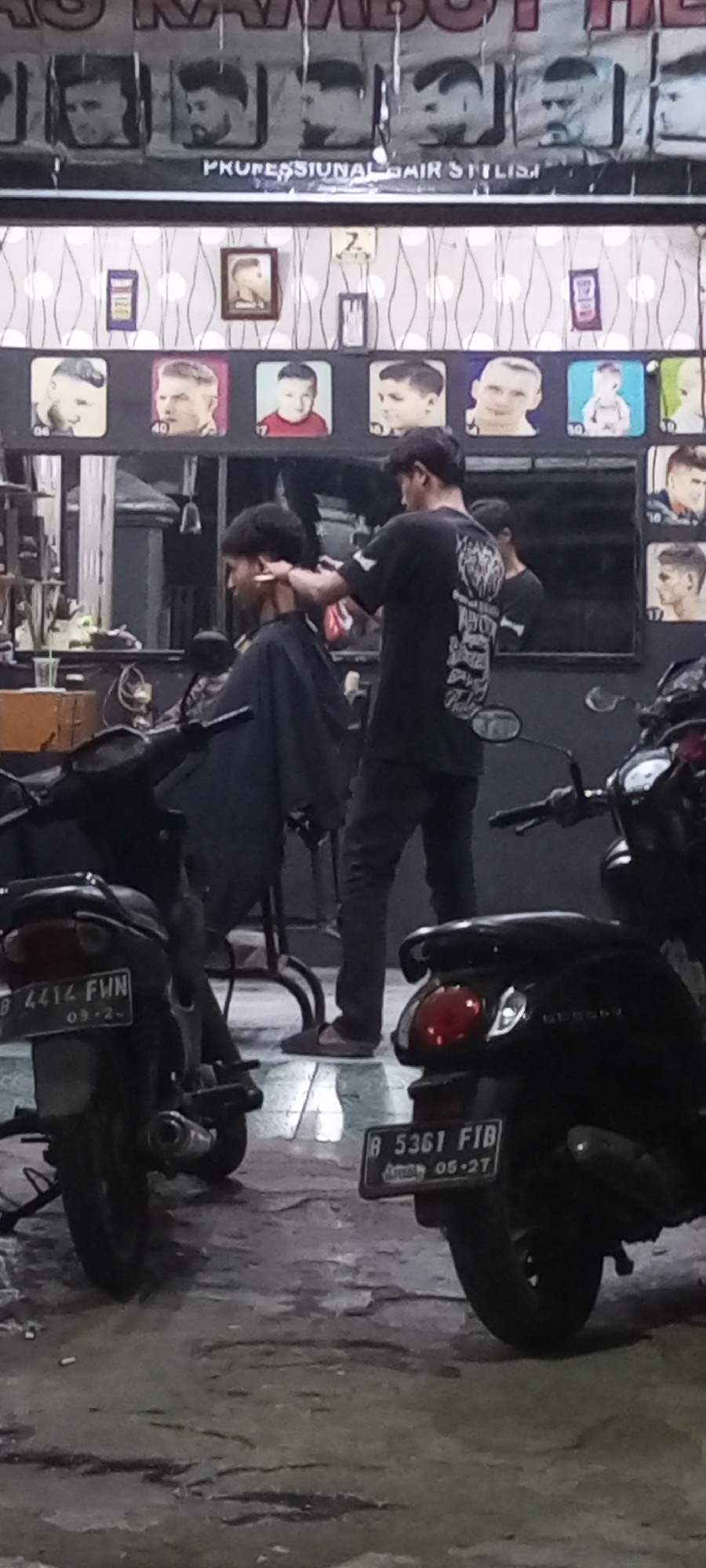 The image depicts the exterior of a barbershop in Setu, Indonesia. In the foreground, two motorcycles are parked on a wet, uneven ground surface. The motorcycle on the left has a license plate reading "B 4414 FWN". The motorcycle on the right has a license plate reading "B 5361 FIB". Behind the motorcycles, inside the dimly lit barbershop, a barber is cutting the hair of a customer. The barber is wearing a black t-shirt with a graphic design on the back and is using clippers. The customer is wearing a black cape. The back wall of the barbershop features framed pictures of various hairstyles, with a sign overhead reading "PROFESSIONAL HAIR STYLIST". The overall atmosphere suggests a typical urban street scene in Indonesia, with the barbershop being an active business.