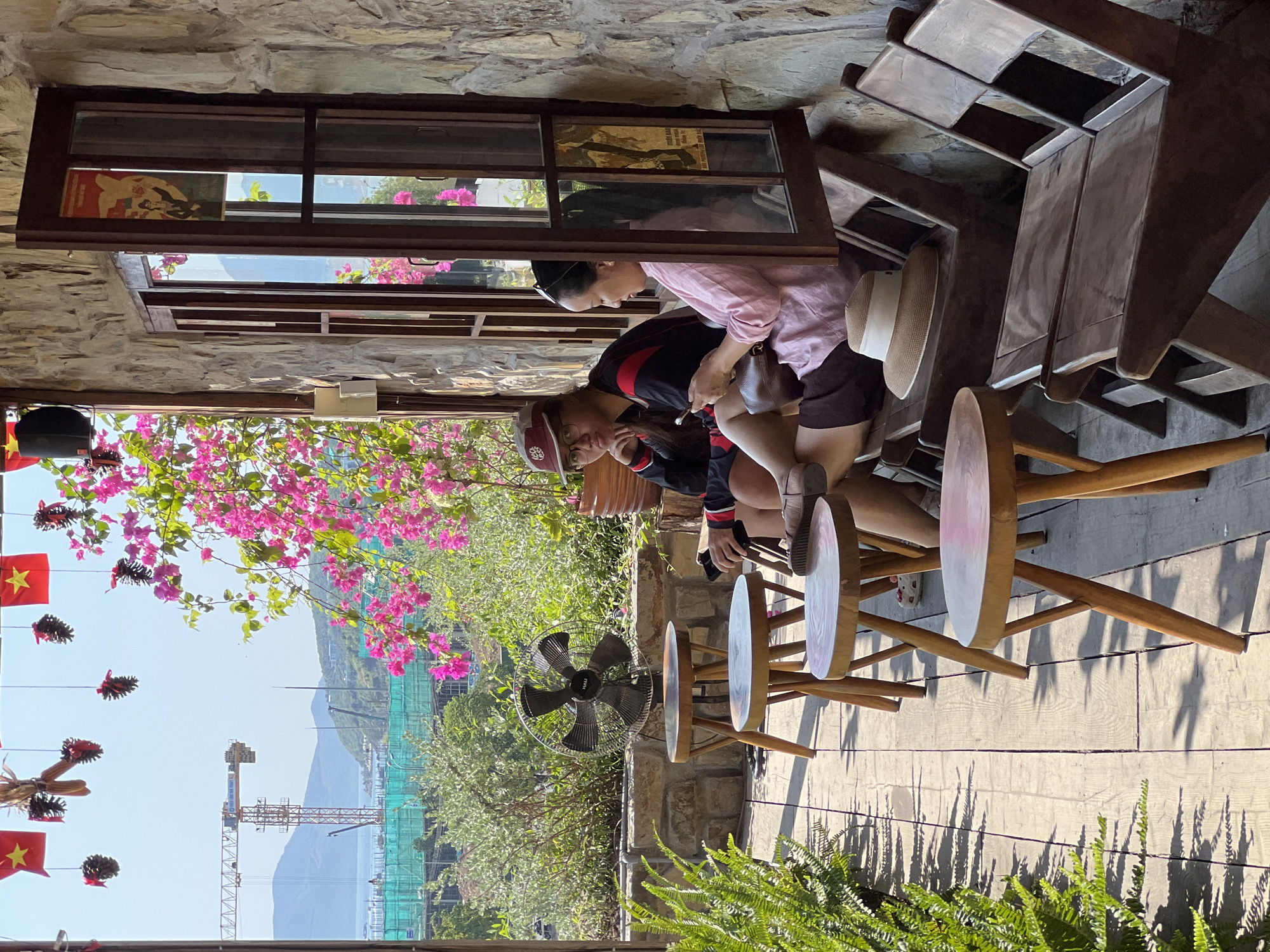 Two women are seated on a wooden bench on a sunlit outdoor patio in Nam Nha Trang Ward, Vietnam. The woman on the left wears a baseball cap, glasses, and a black and red striped long-sleeved shirt with dark shorts. The woman on the right wears a light pink shirt and dark shorts, holding a smartphone. In front of them are several small, round wooden tables. To the left, a black oscillating fan is positioned behind a stone wall. Lush green ferns and flowering pink bougainvillea are visible. In the background, a construction crane and ongoing construction are apparent, along with hills and a body of water. The patio features wooden floorboards and a stone wall. A woven hat rests on the bench between the two women.