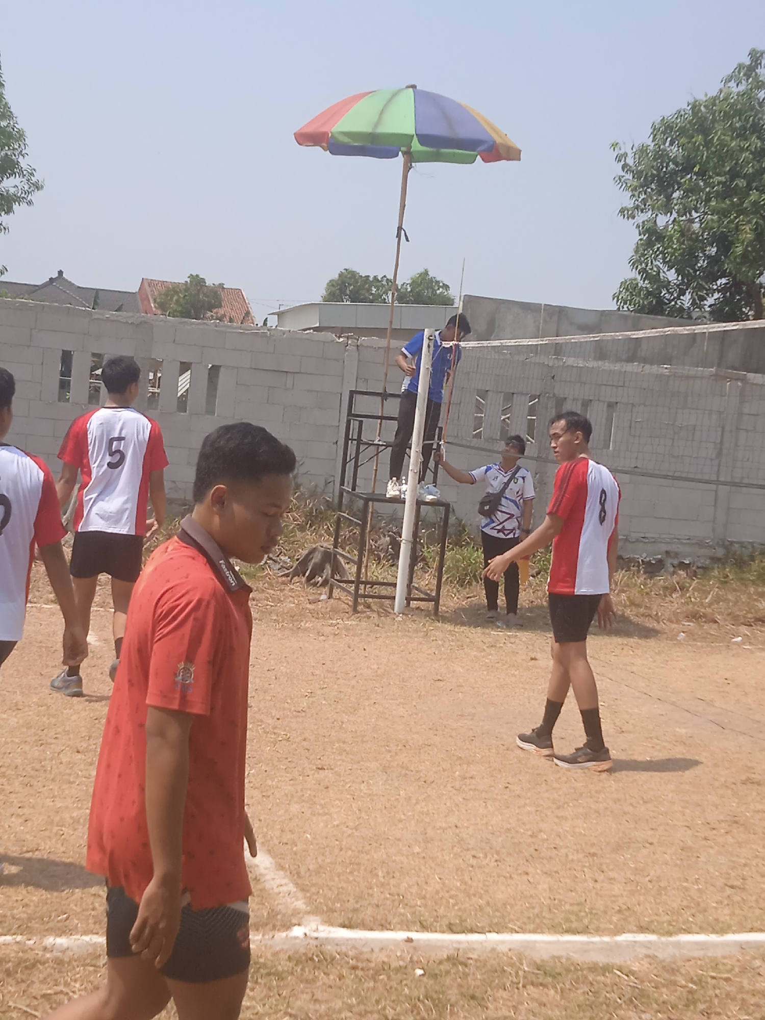 The image depicts an outdoor volleyball court in Klodran, Indonesia, characterized by a dusty brown surface with white boundary lines. A group of young men, presumably participants in a volleyball match, are present. In the foreground, a young man in a red shirt with a patterned design and black shorts is walking away from the camera, his face partially visible. To his left, another individual in a red and white jersey with the number 5 on the back is also walking. In the mid-ground, a young man in a red and white jersey with the number 8 on the back is facing the camera, wearing black shorts and dark socks. Behind him, another person in a white shirt with a colorful geometric pattern and black pants is standing near a volleyball net. Further back, a person in a blue shirt is standing on a raised platform next to the net, holding onto a pole. Above the court, a multi-colored umbrella, supported by a wooden pole, provides shade. In the background, a low concrete wall separates the court from a residential area with buildings and trees visible under a clear, bright sky. The overall scene suggests a casual or amateur volleyball game taking place on a sunny day.