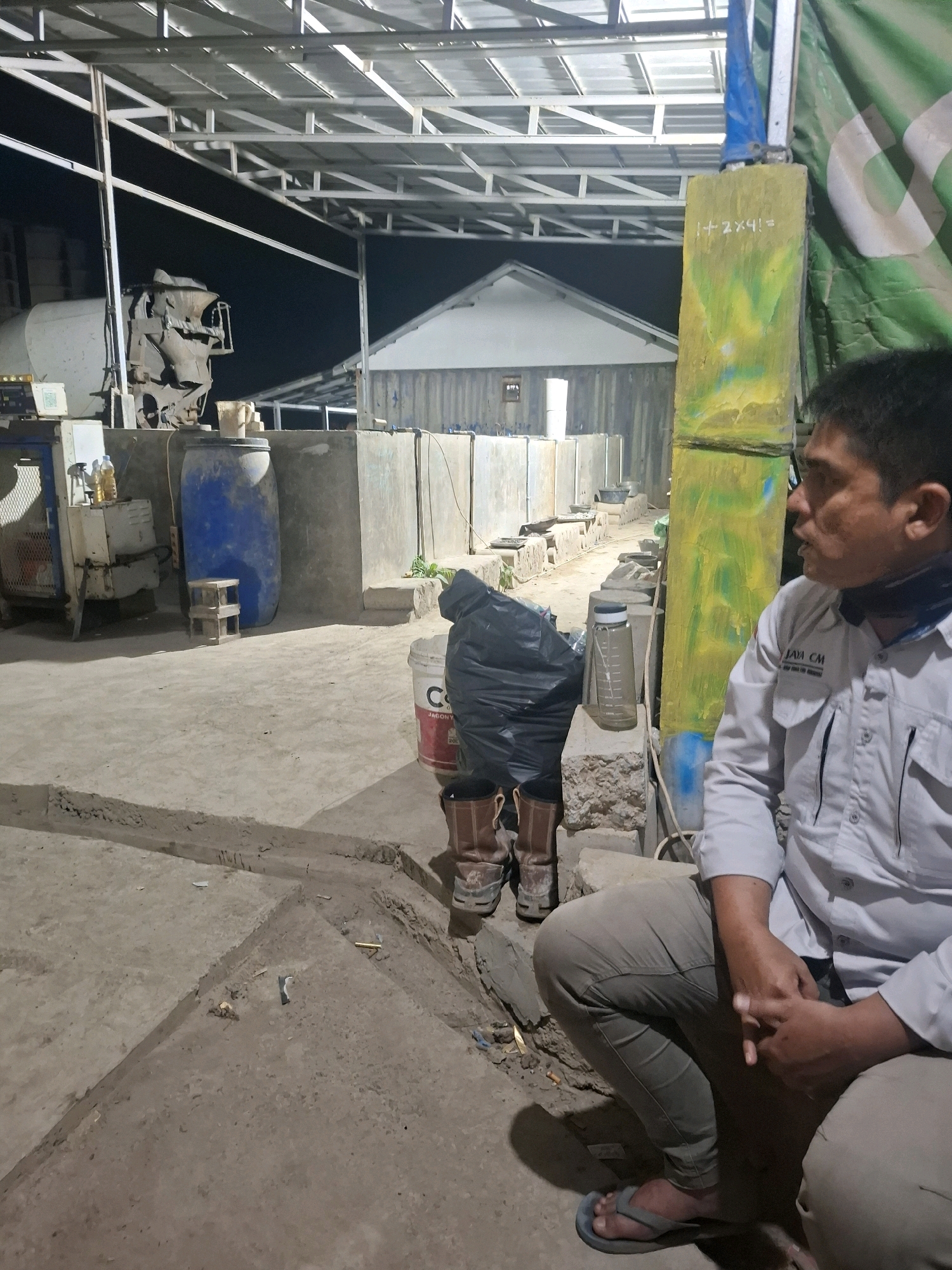 The image depicts an outdoor scene at night, possibly an industrial or agricultural facility, in Kutasirna, Indonesia. A man, wearing a light-colored collared shirt and grey pants, is partially visible on the right side of the frame, crouching. He is looking to his right, with his hands clasped in his lap. His attire suggests he might be a worker at the location.
The environment is characterized by concrete flooring and structures. In the background, a white building with a prominent roofline is visible. Various objects are scattered around, including large blue and white barrels, construction materials, and what appears to be a piece of machinery. A tall, yellow-painted post with some writing, possibly a mathematical equation "1+2X4=", stands prominently near the center. The overall impression is one of a utilitarian, perhaps under-construction or operational site, with minimal lighting contributing to the nighttime atmosphere. The ground is uneven, with trenches or channels carved into the concrete. A pair of brown work boots are visible in the foreground to the left of the man.