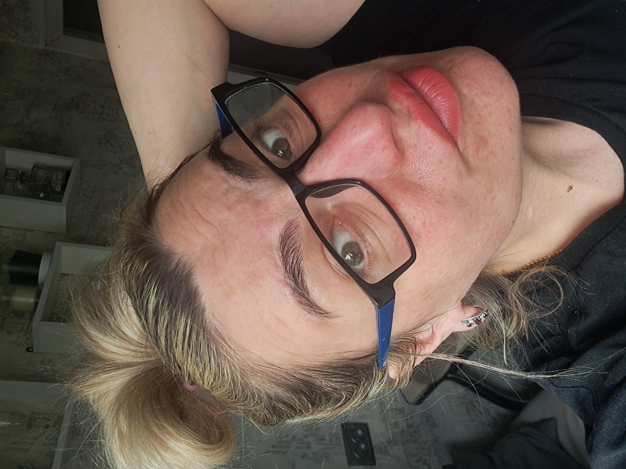 A close-up indoor selfie depicts an adult female subject in Elektrostal, Russia. The subject has light brown and blonde hair styled in a messy bun, with visible darker roots. She wears black-framed glasses with blue temple accents and looks directly at the camera with a wide-eyed expression. Her complexion exhibits some redness and forehead wrinkles. Her lips are painted a coral shade. A silver stud piercing is visible on her left cheek, and a small mole is present on her neck. She wears a dark-colored top, a silver-colored hoop earring with red stones on her visible left ear, and a thin gold-colored chain necklace. Her left arm is raised behind her head, with the elbow partially visible on the upper right side of the frame. The background consists of a grey, textured wall. A dark-colored double electrical outlet is visible on the wall to the left of the subject's head. White open shelving is present in the upper left background, holding a dark cylindrical object, a lighter object, and a framed item.