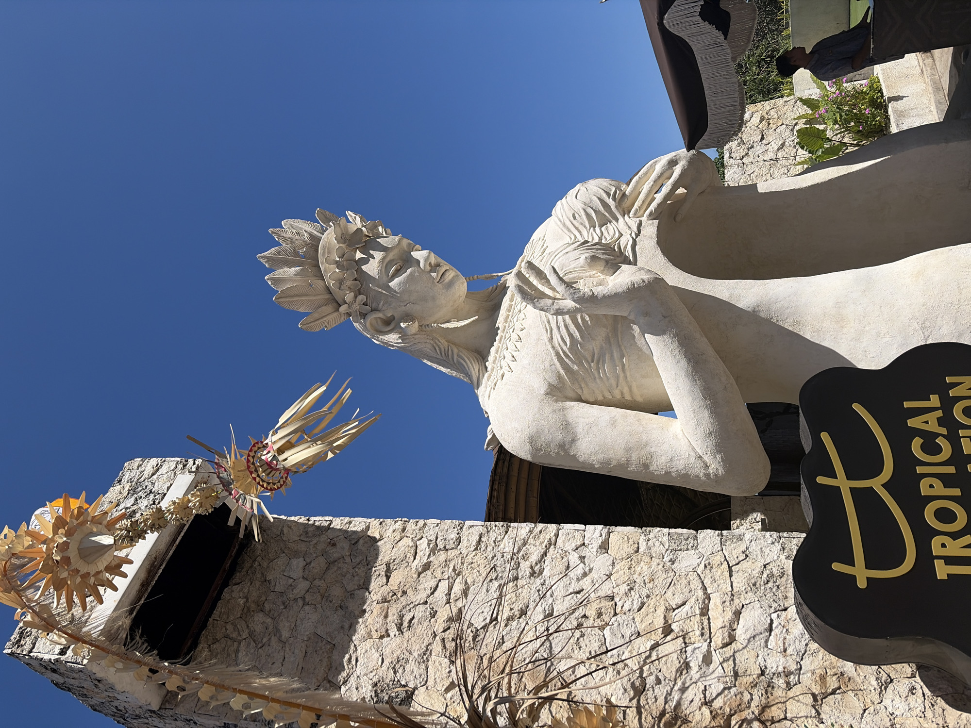 This image captures a stone statue of a woman in Kuta Selatan, Indonesia, set against a bright blue sky. The statue, which appears to be made of a light-colored stone, is positioned to the right of a stone building. It is adorned with a headdress resembling feathers and flowers, and its hands are clasped in front of its chest. Decorative Balinese offerings, made of dried palm leaves and straw, are visible hanging from the stone building. In the foreground, a dark sign with a golden "tt" logo and the word "TROPICAL" is partially visible. To the right of the statue, a person is seated under a brown umbrella with fringe, looking to the side. The environment suggests a warm, sunny day in a tropical location.