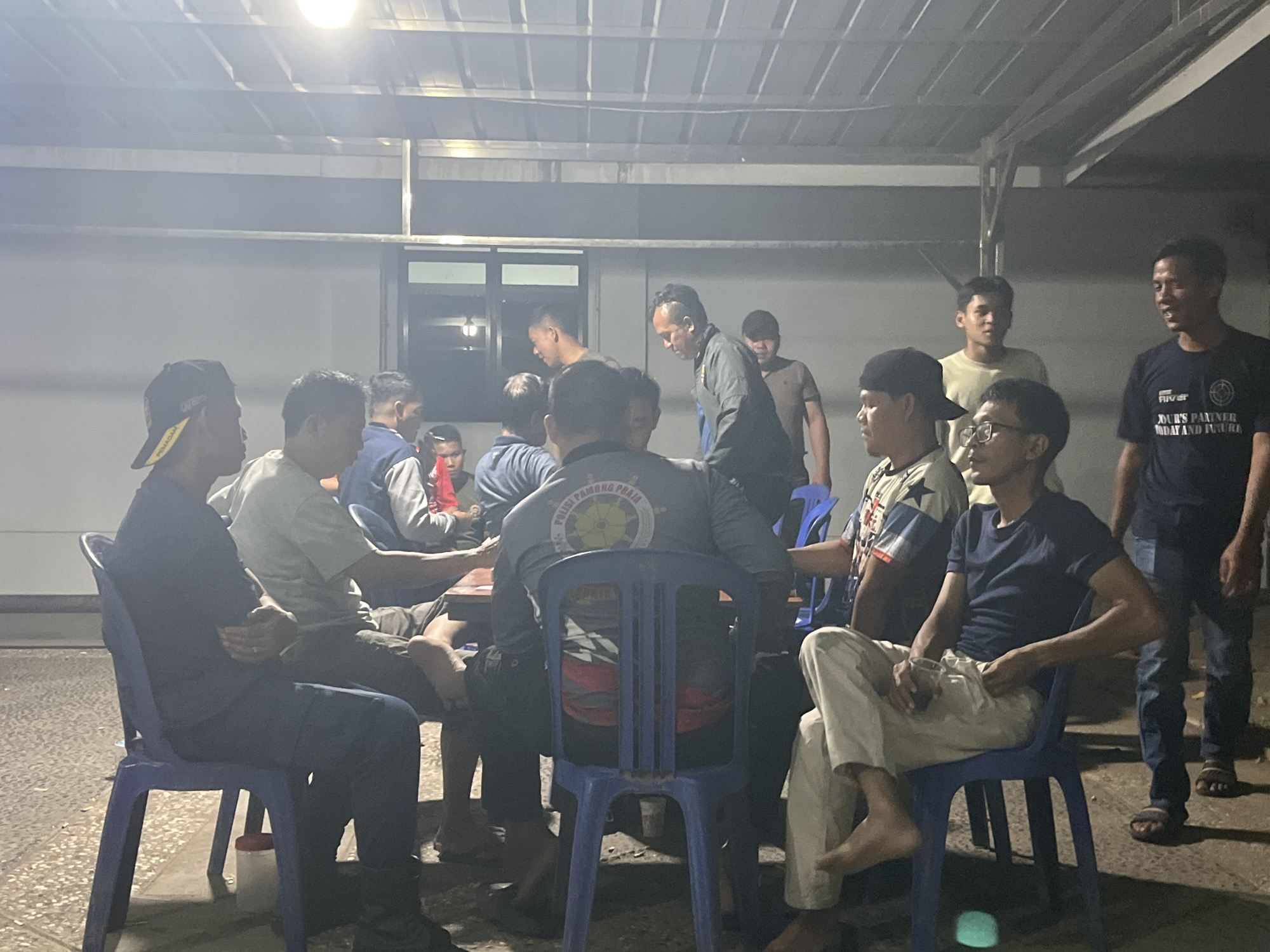 The image depicts a group of men gathered outdoors under a covered structure, likely in Sekayu, Indonesia. The scene appears to be evening or nighttime, with artificial lighting illuminating the area. The men are casually dressed, with some wearing t-shirts and hats, while others are in more formal attire. They are seated on blue plastic chairs around tables, engaged in conversation or possibly a game. One man on the far right is standing and speaking. The environment includes a tiled floor and a plain wall in the background. The overall atmosphere seems relaxed and informal.