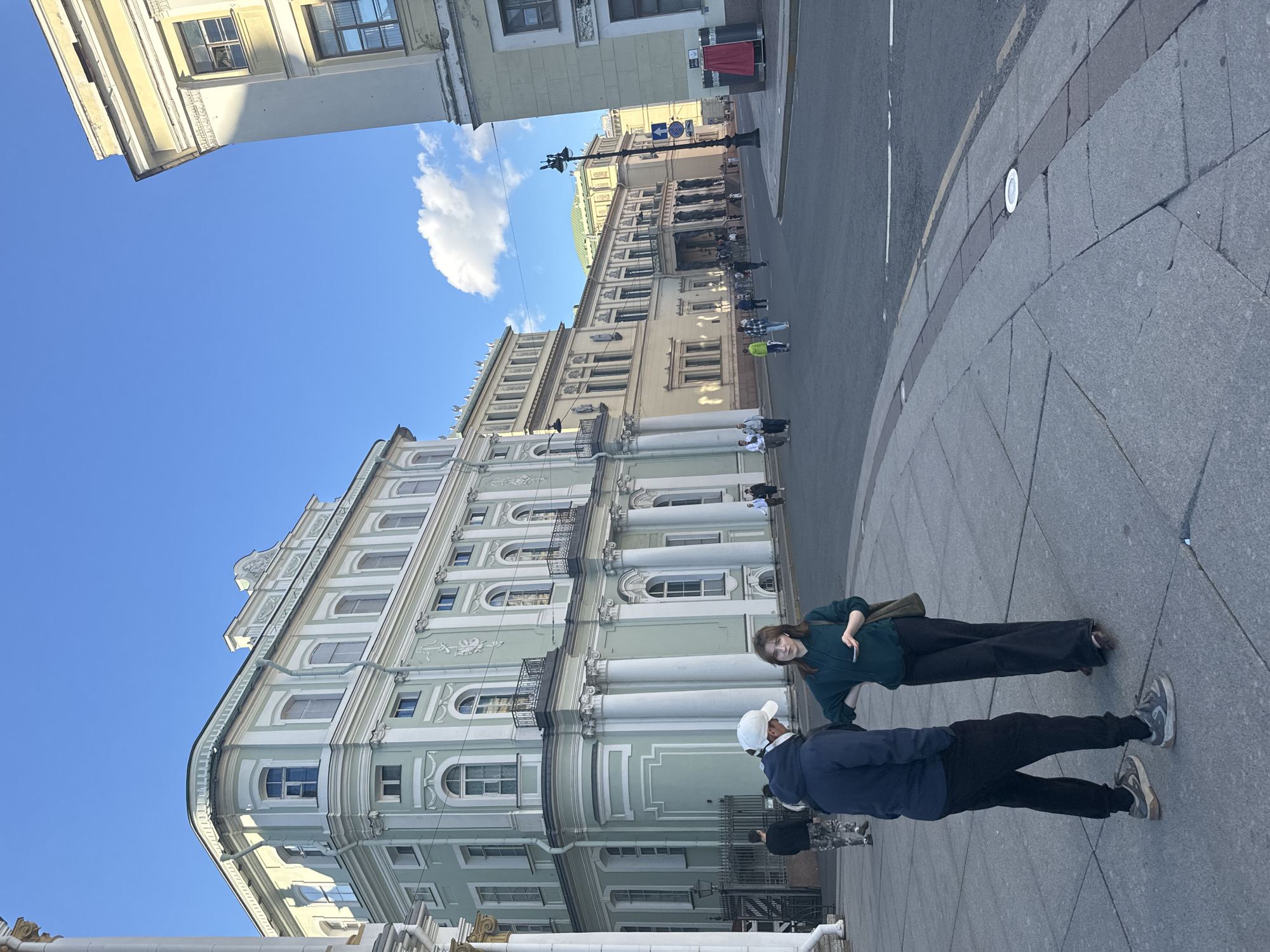 The image captures a street scene in Saint Petersburg, Russia, likely in the area of Palace Square, given the architectural style of the buildings. In the foreground, two individuals, a man and a woman, are engaged in conversation. The man is wearing a dark blue hooded jacket, dark pants, and a white baseball cap. The woman is wearing a dark green hooded sweatshirt and dark pants. They are standing on a paved sidewalk with a curved edge bordering a street.
Behind them, a light green neoclassical building with white decorative elements and columns occupies a significant portion of the frame. This building features multiple windows and balconies. Adjacent to it, another long, pale yellow building with a similar architectural style extends further down the street. The street itself is asphalt, with painted lane markings.
Several other people are visible in the middle ground, walking along the street and sidewalk. The sky is bright blue with a few scattered clouds. The overall impression is a sunny day in an urban environment with grand historical architecture.