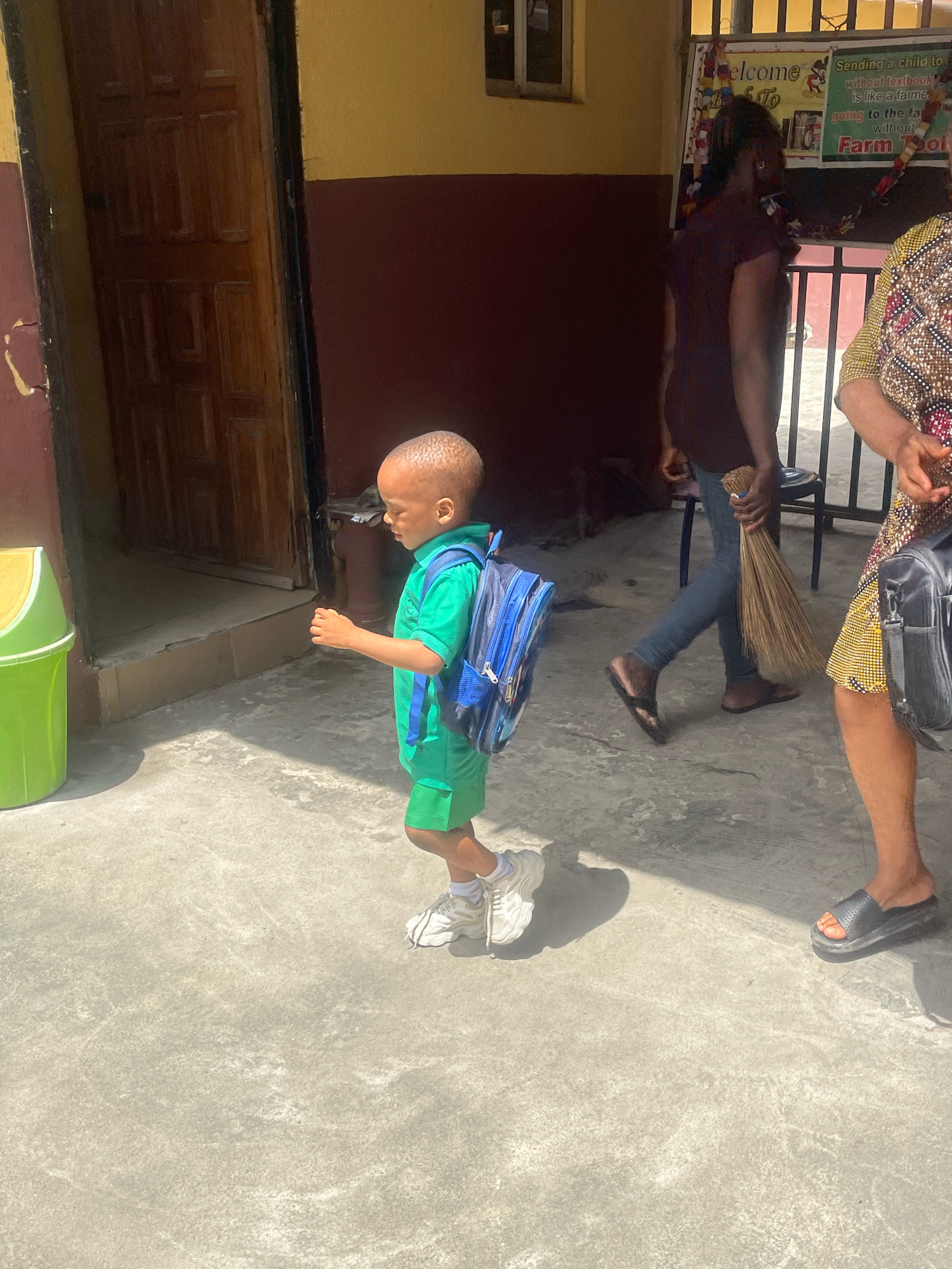 A young boy, with short, close-cropped hair, wearing a bright green t-shirt and shorts, and white sneakers, walks from right to left across a concrete surface. He has a blue backpack on his back. To his left, a green plastic trash can with a yellow lid is visible. Behind the boy, a dark wooden door is ajar, leading into a building with a yellow and dark red exterior. In the background, a woman in a dark shirt and jeans walks from left to right, holding a broom. To the far right, a woman wearing a patterned dress walks away from the camera, carrying a black bag. There is a sign on a bulletin board in the background with text, including "Welcome" and "Farm School." The scene appears to be outdoors, possibly in the courtyard of a school in Ado, Nigeria.