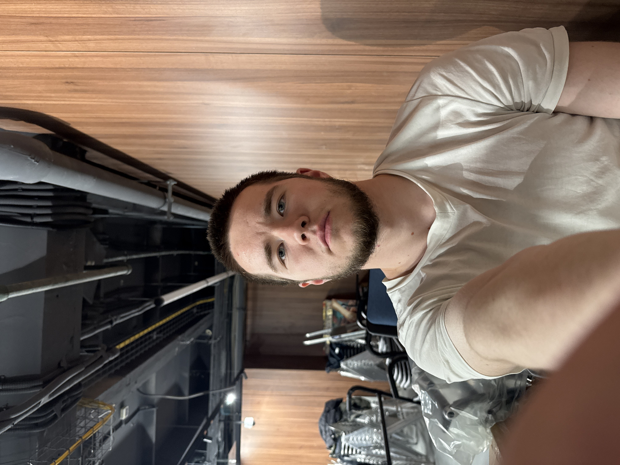 A young man with short dark hair and a beard is the primary subject of the image, which appears to be a selfie. He is wearing a white t-shirt. The background shows what appears to be a storage or utility area with exposed pipes, wiring, and metal scaffolding. There are stacks of metal chairs covered in plastic wrapping. Wooden paneling is also visible. There are no clear indicators of the specific location in Kazan, Russia, or any interactions taking place.