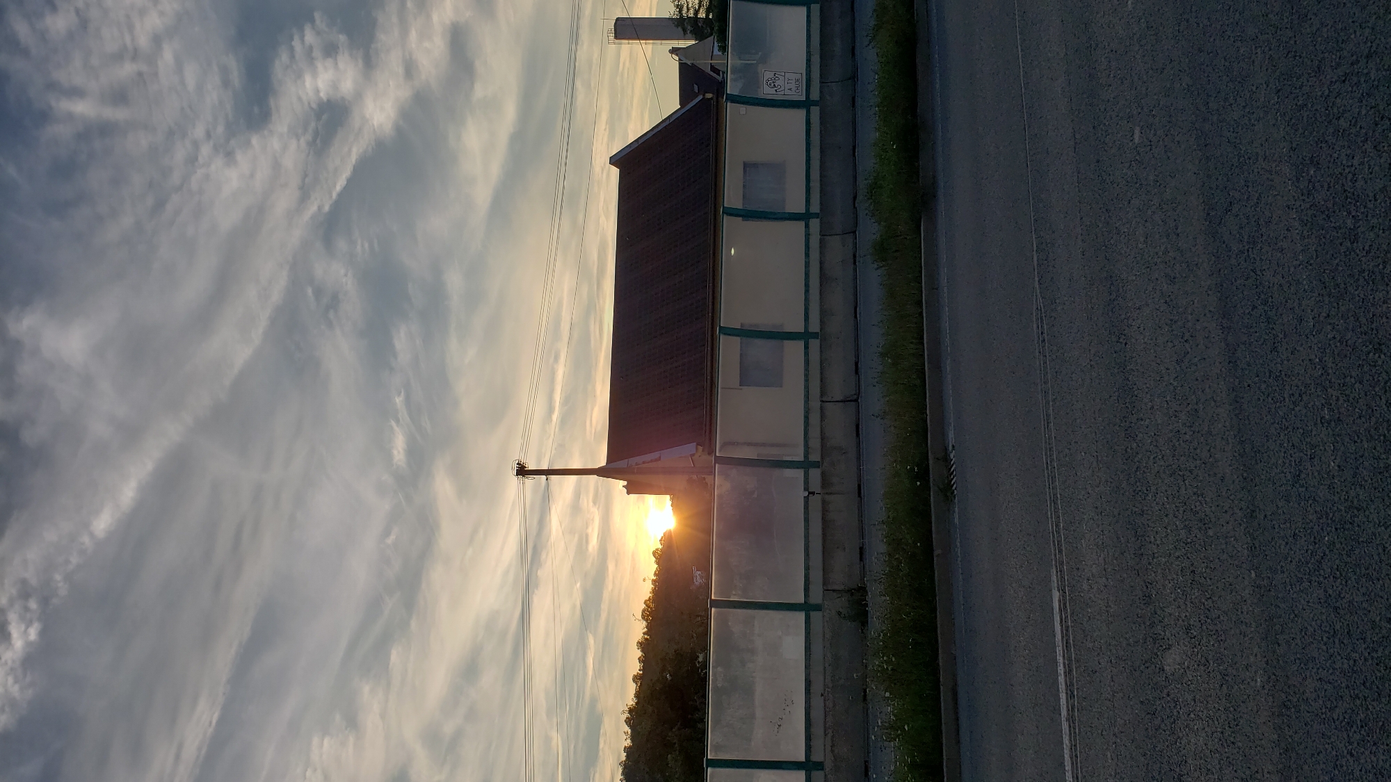 The image depicts a sunset in Prešov, Slovakia. The sun is setting behind a building with a dark roof, casting a warm glow. In the foreground, there is a road with a concrete barrier and a sound barrier. Trees are visible in the background, and the sky is filled with wispy clouds. The scene appears to be a quiet evening on the outskirts of the city.