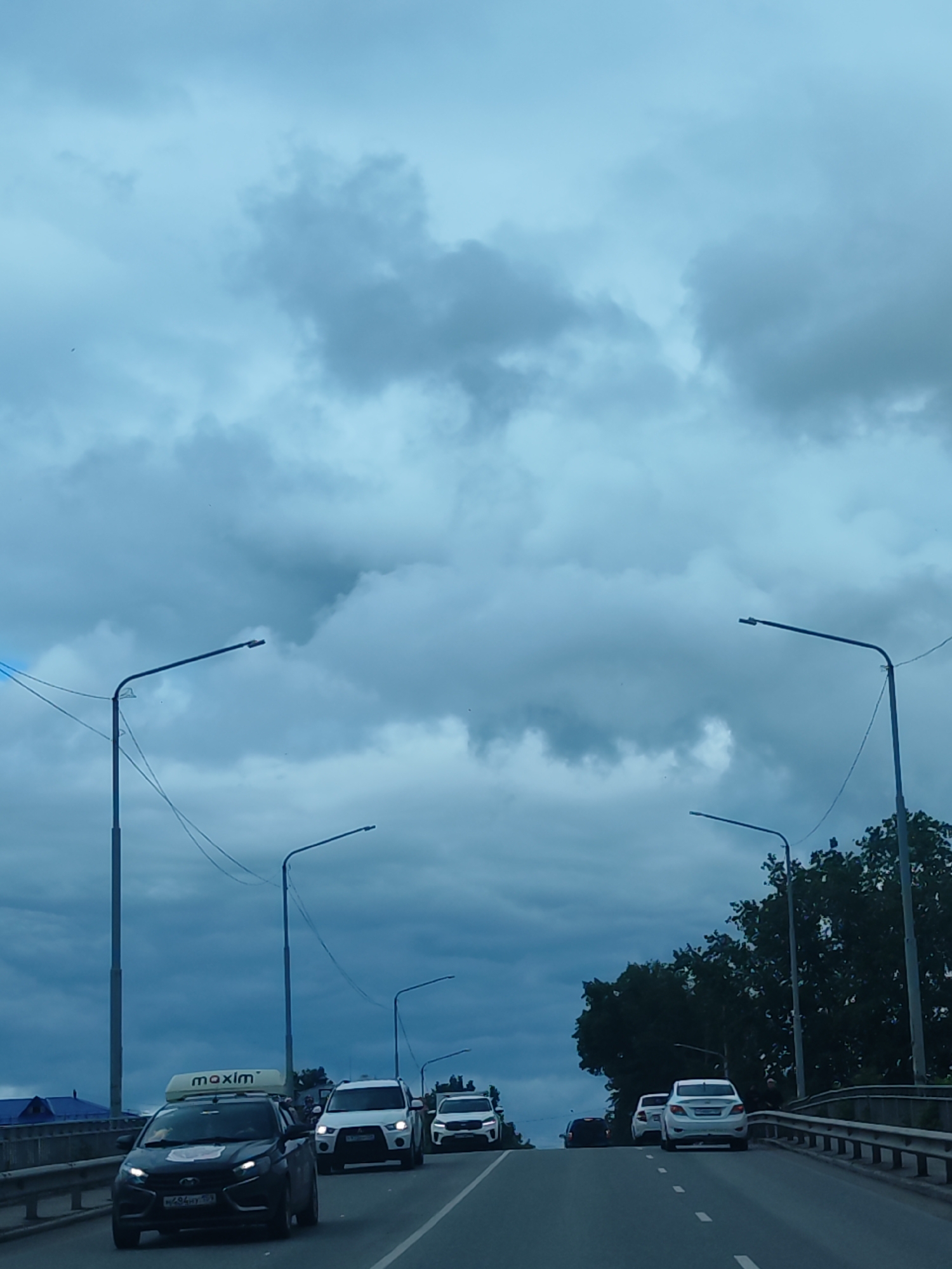 The image shows a view from a car traveling on a bridge in what appears to be the city of Chaikovsky, Russia. The sky is overcast with heavy clouds. Streetlights line the sides of the bridge. Traffic includes several vehicles, such as a dark-colored car with a "maxim" sign on top, a white SUV, and other passenger cars. The road is marked with white lines. Vegetation is visible on the right side of the frame, with trees beyond the bridge railing. A building with a blue roof can be spotted in the background.