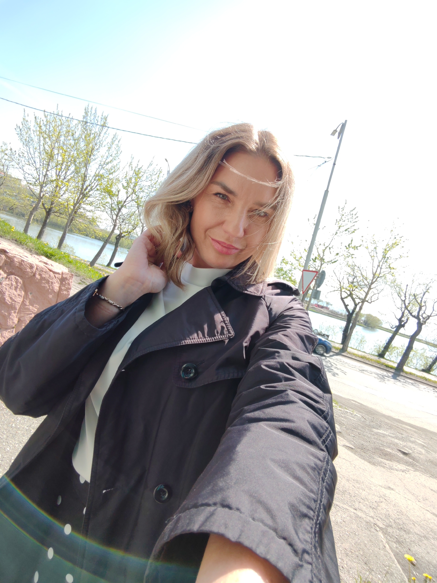 An outdoor, eye-level, medium shot shows a fair-skinned woman in her early 40s with blonde hair and blue eyes, taking a selfie. She is wearing a black trench coat over a white top. Her left hand is touching her hair, and her right arm is extended towards the camera. She is smiling and looking directly at the viewer. The background consists of a body of water, trees, and a street with a lamppost and a traffic sign. The sky is clear and blue.