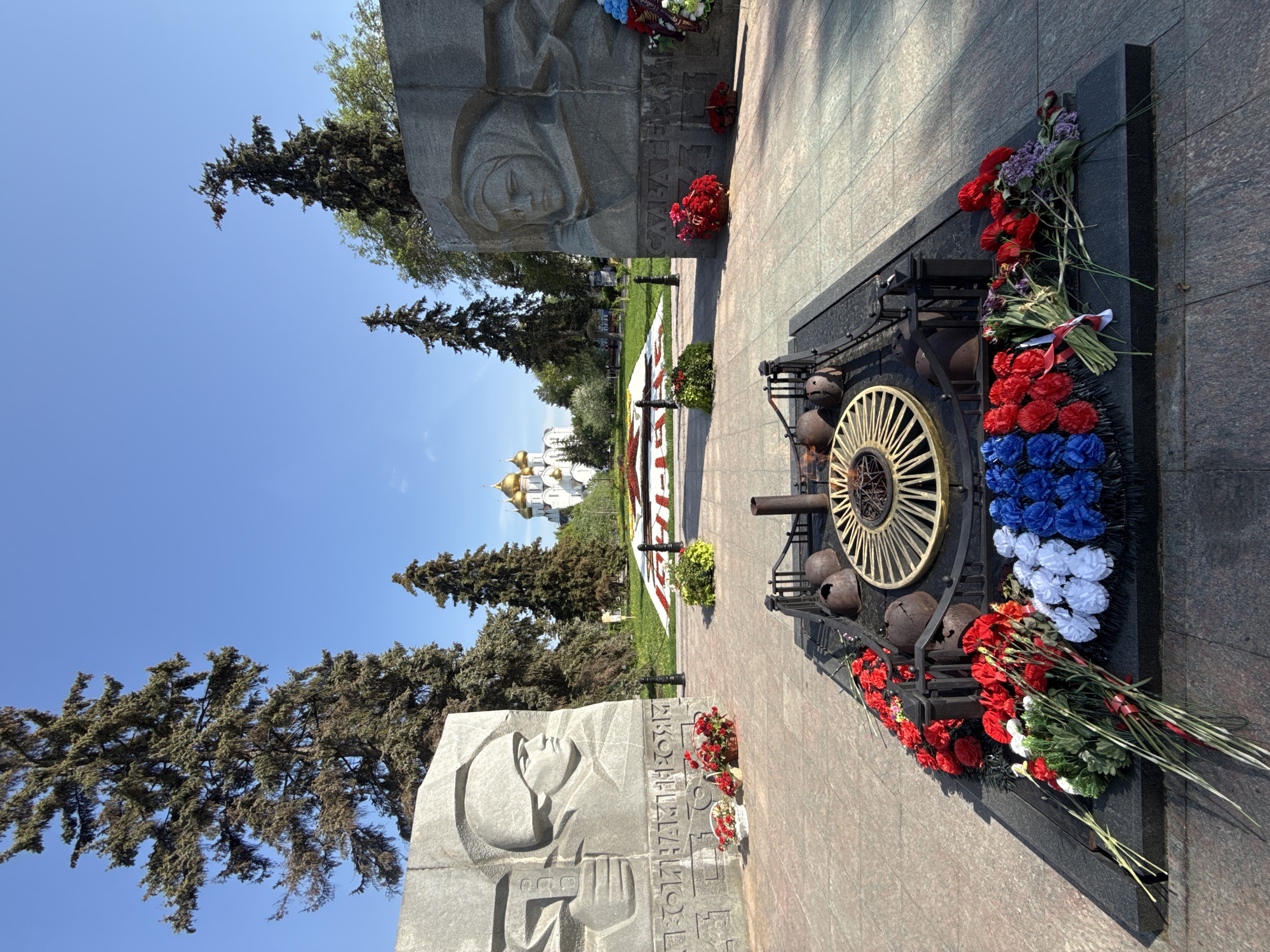 The image shows a war memorial in Rusanovo, Russia, consisting of two grey stone sculptures depicting human faces, potentially soldiers or civilians. Between them is a black granite structure adorned with a golden wheel-like element and spent artillery shells. Flowers, arranged in the colors of the Russian flag (red, white, blue), as well as bouquets of red flowers, are placed on the memorial. In the background, a Russian Orthodox church with golden domes is visible. The sky is clear and blue, and tall green trees are present on both sides of the memorial. The ground is paved with red-brown stone.