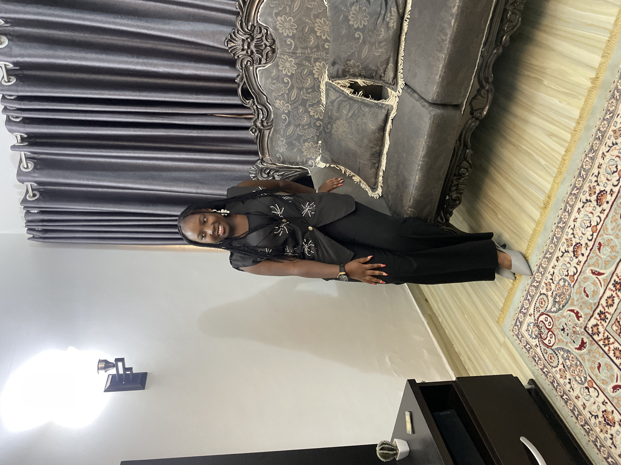 The image, taken in Jos, Nigeria, features a young woman standing and smiling towards the camera in what appears to be an indoor living space. She has dark braided hair and is wearing a chic black outfit consisting of a short-sleeved top adorned with sparkling, star-like embellishments and black trousers. She also sports a dark wristwatch, light-colored earrings, and light grey pointed-toe flats. Her nails are painted a reddish-orange hue.
Behind her, to her left, is a large, ornate sofa with a dark grey, patterned fabric and decorative carved wooden elements on its frame. Several fringed cushions are placed on the sofa. Grey pleated curtains with silver grommets hang along the back wall, suggesting a window.
The floor on the right side of the image is covered by a light-colored rug featuring intricate patterns in shades of light blue, beige, brown, and white, with fringed edges. A light wooden or laminate floor is visible next to the rug.
On the white wall to her right, a modern-style wall sconce with a bright, glowing bulb illuminates the area. In the bottom right corner, a dark brown wooden cabinet or drawer unit is visible, with a small succulent plant in a white pot placed on top. The overall lighting suggests a well-lit interior, possibly during the day. The woman appears happy and relaxed, posing for the photograph. No visible text is present in the image.