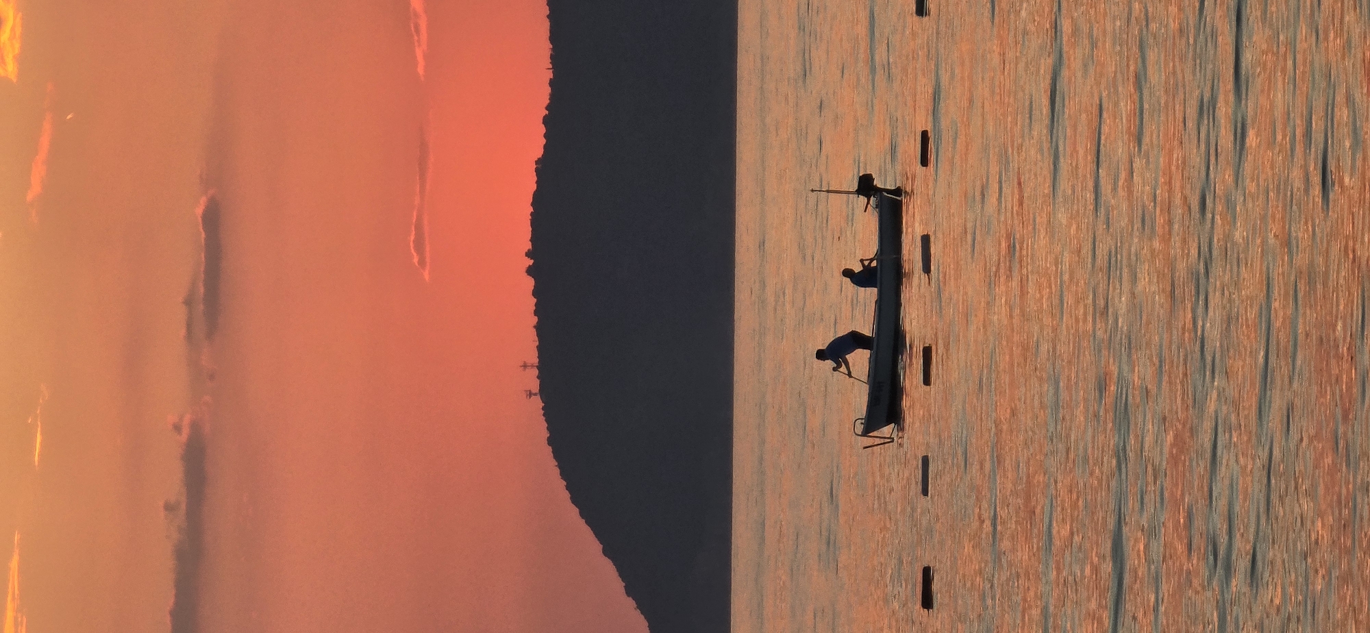 The image shows a horizontal shot of a sunset scene with a boat on the water. The sunset fills the left side of the frame with warm orange and pink colors. A dark mountain range is silhouetted against the sunset. On the right side of the frame is water with reflections of the sunset. A long, narrow boat with three people on board is centered in the water. The first person at the front of the boat is bent over and appears to be rowing with a pole. The second person is sitting in the middle of the boat, and a third person is standing at the back. There are also some wooden objects or posts sticking out of the water near the boat. The location is the city of Bar, Montenegro, and the time of day is sunset.