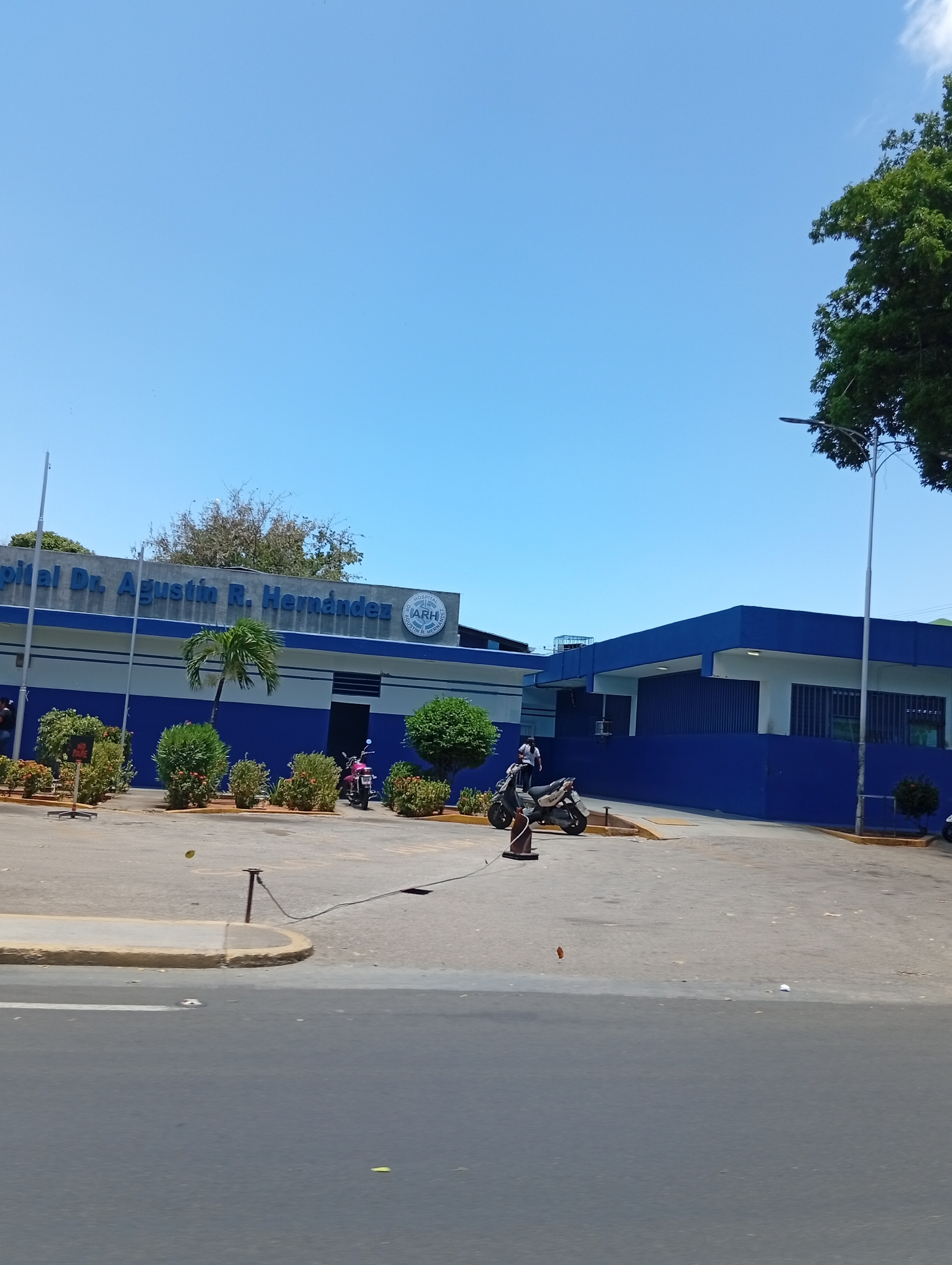 The image shows the Hospital Dr. Agustín R. Hernández in Juan Griego, Venezuela. The hospital building is blue and white with the hospital's name clearly displayed on the facade. In front of the building, there is a paved area with landscaping, including trees and bushes. Several motorcycles are parked in the parking area. A curb separates the parking area from the street. A metal post with a wire attached to it is located near the curb. The sky is clear and blue, indicating a sunny day. A tall tree is visible on the right side of the frame.