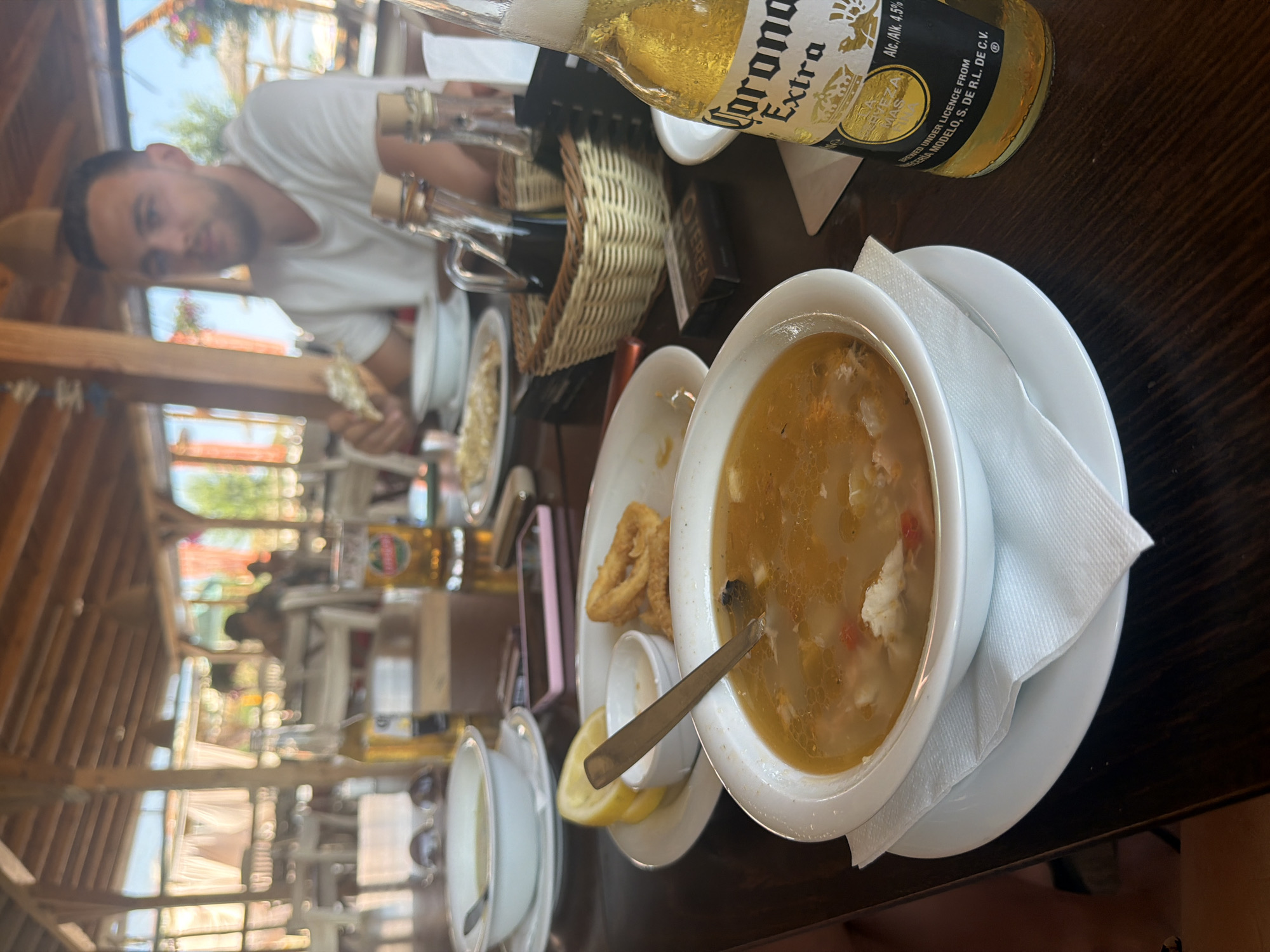 This image depicts a dining scene at a restaurant in Sinemorets, Bulgaria, likely on a sunny day. The foreground features a wooden table with various dishes and drinks. A white bowl containing a hearty soup with visible chunks of seafood and vegetables is centrally placed, with a spoon resting inside. To the right, a bottle of Corona Extra beer with its distinctive label is positioned, partially obscuring a woven basket filled with condiments. A small white dish containing what appears to be a dipping sauce and a lemon wedge are on a white plate beside the soup. Further back, another white bowl and plate are visible. In the background, a man in a white t-shirt is seated at a table, holding a piece of food and looking towards the right. Other diners and restaurant structures are blurred in the background, suggesting an open-air or partially covered dining area. The ambiance appears relaxed and casual, consistent with a seaside resort town like Sinemorets.