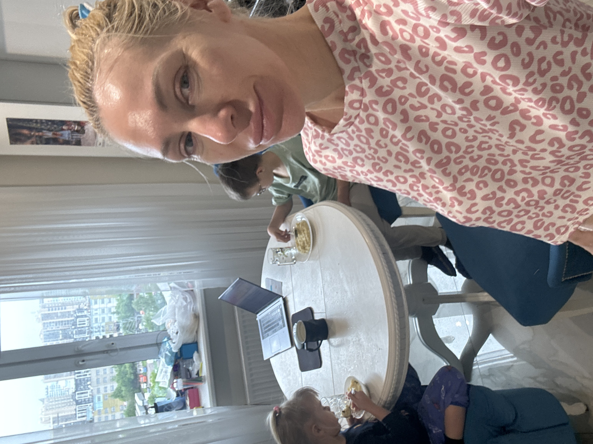 The image shows a woman taking a selfie in an apartment. She has blonde hair pulled back and is wearing a pink and white patterned top. In the background, there is a round white table with a laptop, a mug, and plates of food. Two children, a boy and a girl, are seated at the table, appearing to be eating. A window in the background reveals a cityscape. There are miscellaneous items on the windowsill. The location is Unknown, Unknown.
