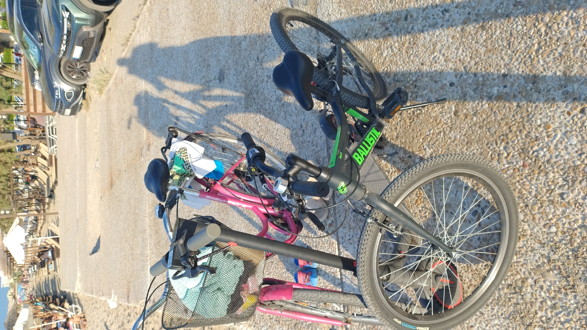 In Perea, Greece, two bicycles are parked on a gravel path adjacent to a sandy beach. The bicycle in the foreground is black and green with the brand "BALLISTIC" visible on its frame. It has a black seat and handlebars, and its front wheel is large with prominent tire treads. Behind it, a pink bicycle is visible, featuring a basket on the handlebars containing a light blue towel and a white and green patterned item. A shadow of a person is cast on the ground to the right of the bicycles. In the background, beach umbrellas and lounge chairs are set up on the sand, with several people relaxing and walking. Two cars, a dark gray SUV and a greenish SUV, are parked further in the background, near a treeline. The overall scene depicts a sunny day at a coastal location.