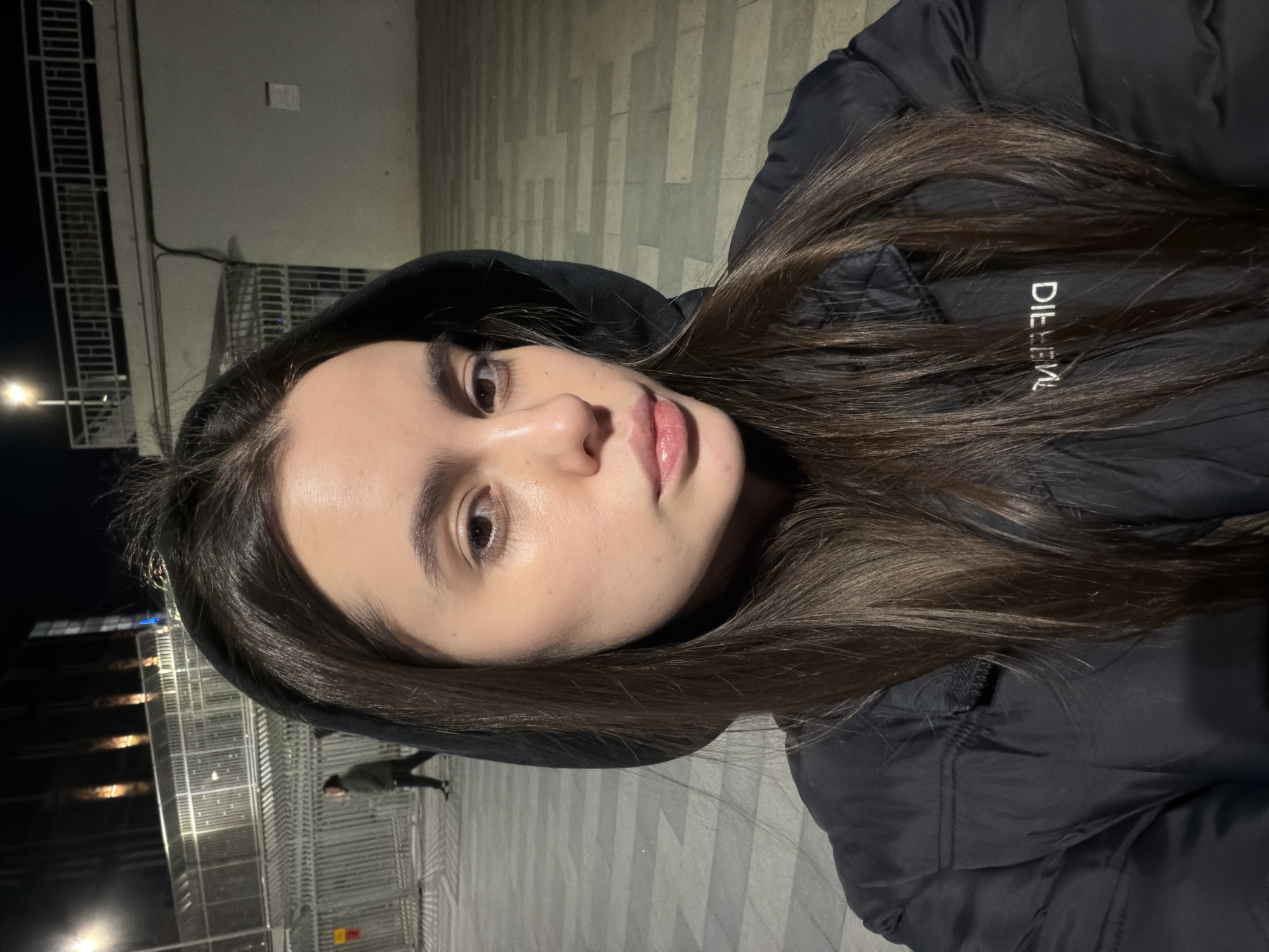 A woman with brown hair is wearing a black hooded jacket. She is outside at night. The background appears to be a public space in the city of Perm, Russia, indicated by the street lamps and the pavement design. A person is walking in the background on the left side of the frame. The jacket has white letters on the side.