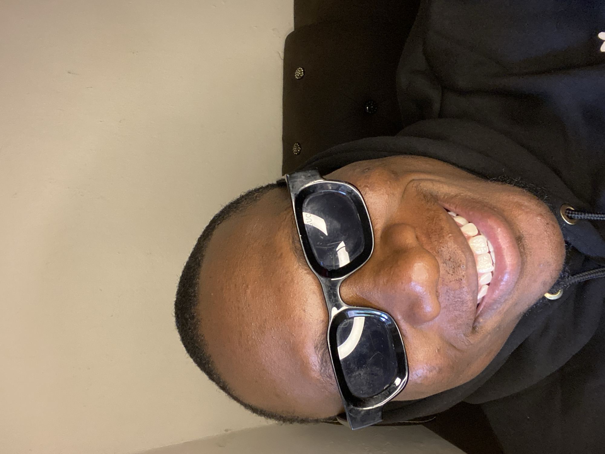 The image presents an angled, close-up portrait of a person with dark skin, appearing to be lying down or reclining, with their head turned to the side. The person is smiling broadly, revealing bright white teeth, conveying an emotion of happiness or confidence. They are wearing black-framed sunglasses with dark lenses; the word "GUCCI" is faintly visible in the reflection of the right lens. The individual is dressed in a black hoodie, with a black drawstring visible near the neck. The background consists of a plain, light-colored wall on the left side of the frame and a dark, possibly upholstered surface with decorative, metallic studs on the right, suggesting an indoor setting. No specific activity is depicted beyond posing for the photograph. This scene is located in Bukuru, Nigeria.