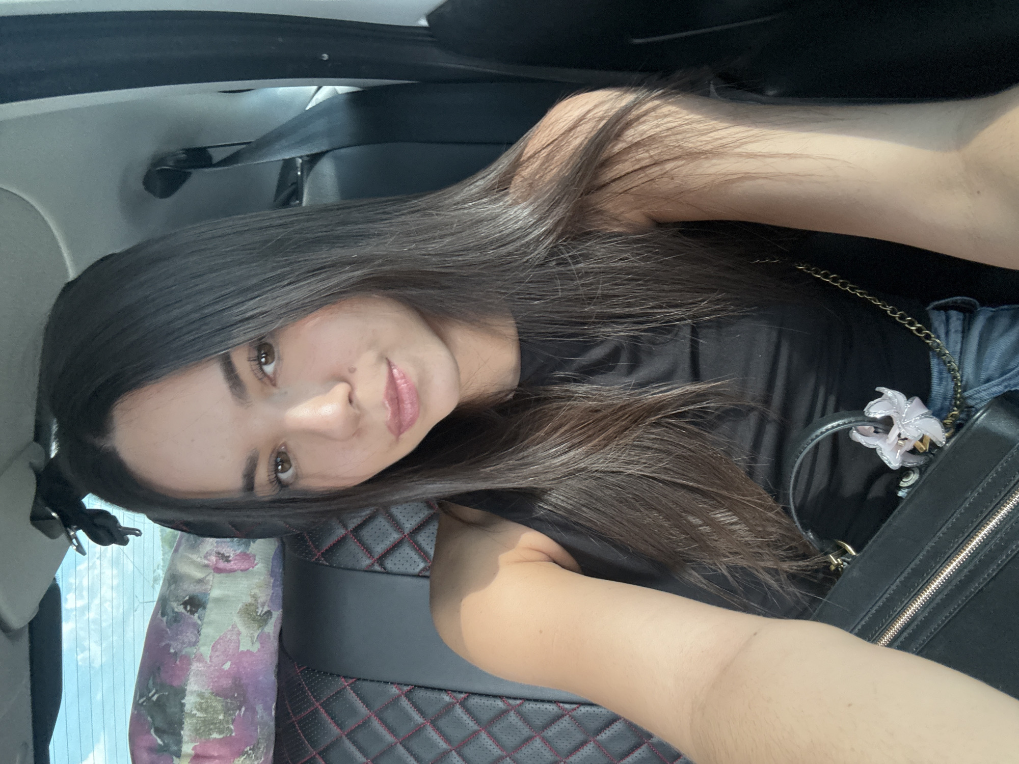A young woman with long, dark hair is sitting in the back seat of a car, taking a selfie. She is wearing a black tank top and blue jeans. A black handbag with a floral charm hangs from her shoulder. The car seat is black with red stitching. A floral pillow is visible in the background, propped against the back window. The window is open, revealing a blurred outdoor scene with a bright sky. The overall lighting suggests it is daytime.