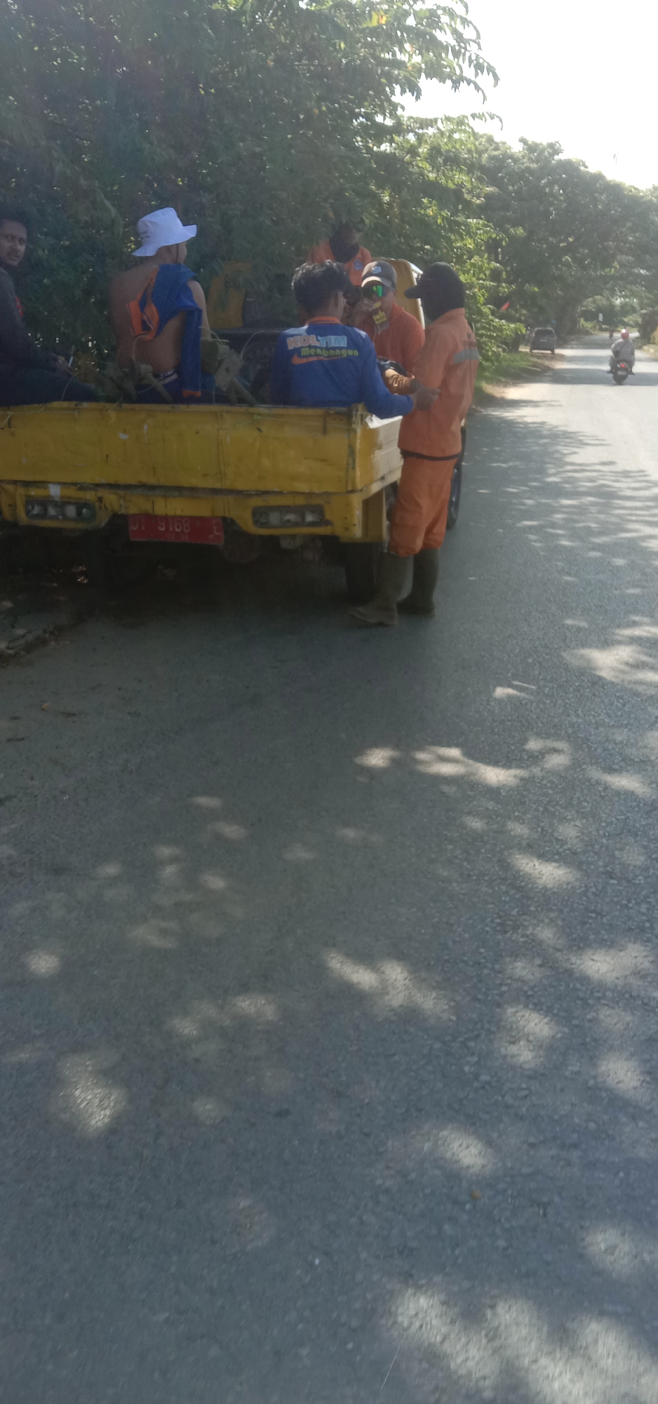 On a sunny day in Kendari, Indonesia, a yellow utility truck is parked on the side of a road. Several individuals are gathered in and around the truck bed, appearing to be workers. One person, wearing a white hat and blue shirt, is seated in the truck. Another, in a blue long-sleeved shirt with "MOLYTIN Mendoagae" printed on it, is also in the truck. Two individuals are dressed in orange work uniforms. One of them is wearing dark boots. In the background, trees line the road, and a motorcycle is visible in the distance. The truck's license plate reads "DT 9168". Shadows from the trees are cast across the asphalt road.