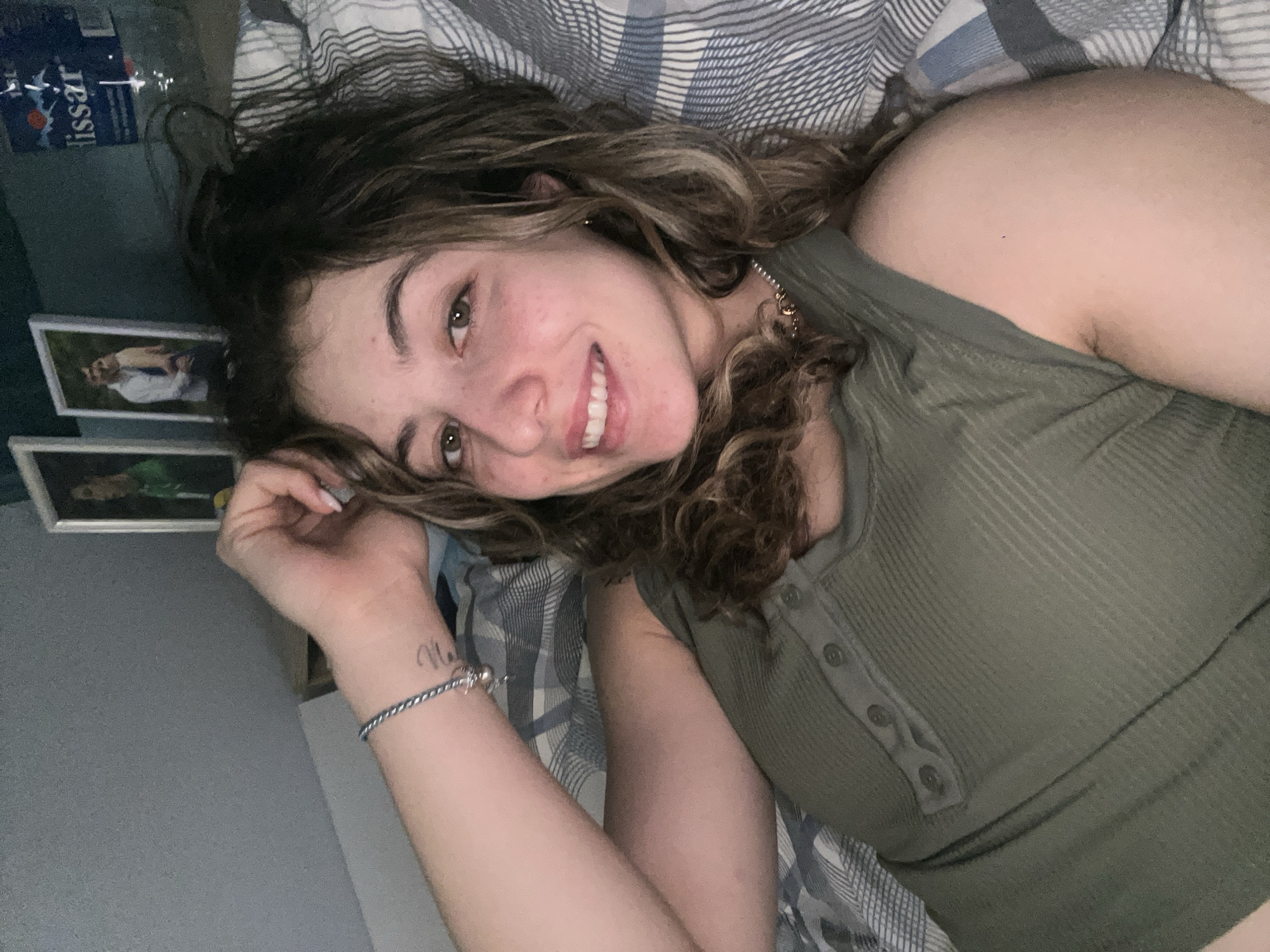A woman with curly brown hair is lying on a bed with a grey and white striped sheet. She is wearing an olive green button-up shirt and a bracelet on her left wrist. The woman is smiling and looking at the camera. Behind her, on a wall, there are two framed pictures and a water bottle. There is no information in the image to suggest this was taken in Plovdiv, Bulgaria.