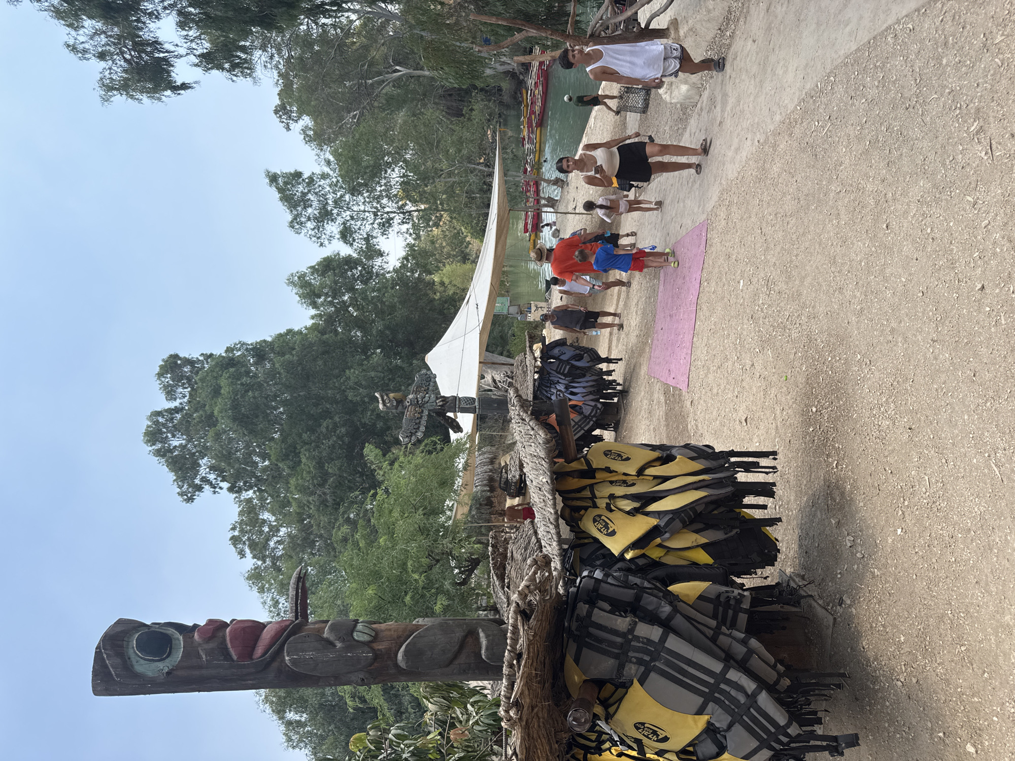 The image depicts an outdoor scene in Emek HaYarden Regional Council, Israel, likely a recreational area near a body of water. In the foreground, numerous yellow and black life jackets are hanging from a wooden structure. A prominent wooden totem pole with carved facial features stands to the left. A pathway, covered in gravel and dirt, leads into the background. Several people are walking on this path. Some are dressed in casual summer attire. In the middle ground, a large white tent provides shade. Beyond the tent, a river or lake is visible, with several red and yellow kayaks lined up on its surface. The riverbanks are lined with lush green trees. The sky is clear and pale blue. The overall atmosphere suggests a warm, sunny day at a water-based attraction or park.