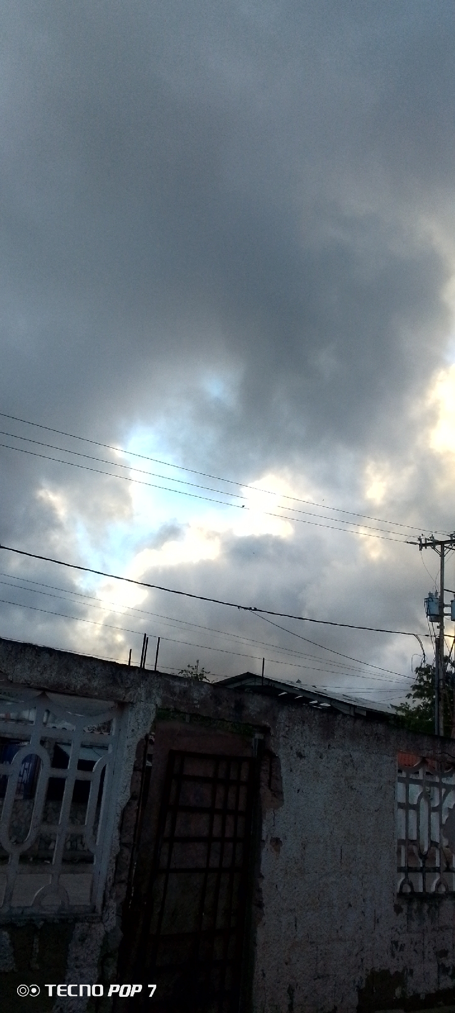 This is a vertical, outdoor, medium shot of a cloudy sky above a residential building's rooftop. The sky is filled with dark, grey clouds, with patches of bright light peeking through in the upper right and center. Several power lines are stretched horizontally across the frame, and a utility pole with transformers is visible on the right side of the image. The rooftop below is constructed of concrete and appears weathered and somewhat damaged in places. On the left, there is a white decorative fence or railing. In the center, a dark, metal-barred gate is set into a rough opening in the wall. The lighting suggests it is either early morning or late afternoon, possibly before or after a storm. There is no discernable activity or emotion depicted. The bottom left corner of the image displays the text "OO TECNO POP 7" in white. The setting is likely a residential area in Altagracia, Venezuela, given the context provided.