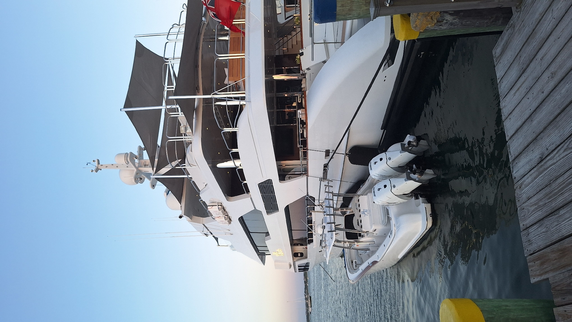 This is a photograph taken on the water in Nantucket, Massachusetts, United States of America. The scene features a large white luxury yacht docked next to a smaller white boat. The yacht has multiple decks, dark-tinted windows, and appears to be quite long. On the upper deck, a dark-colored awning is visible. The smaller boat, which is closer to the viewer, has two white outboard motors and is equipped with fishing rod holders. In the background, a sailboat is visible in the distance. The water is calm with ripples, and the sky is a clear blue. The foreground consists of a wooden dock.