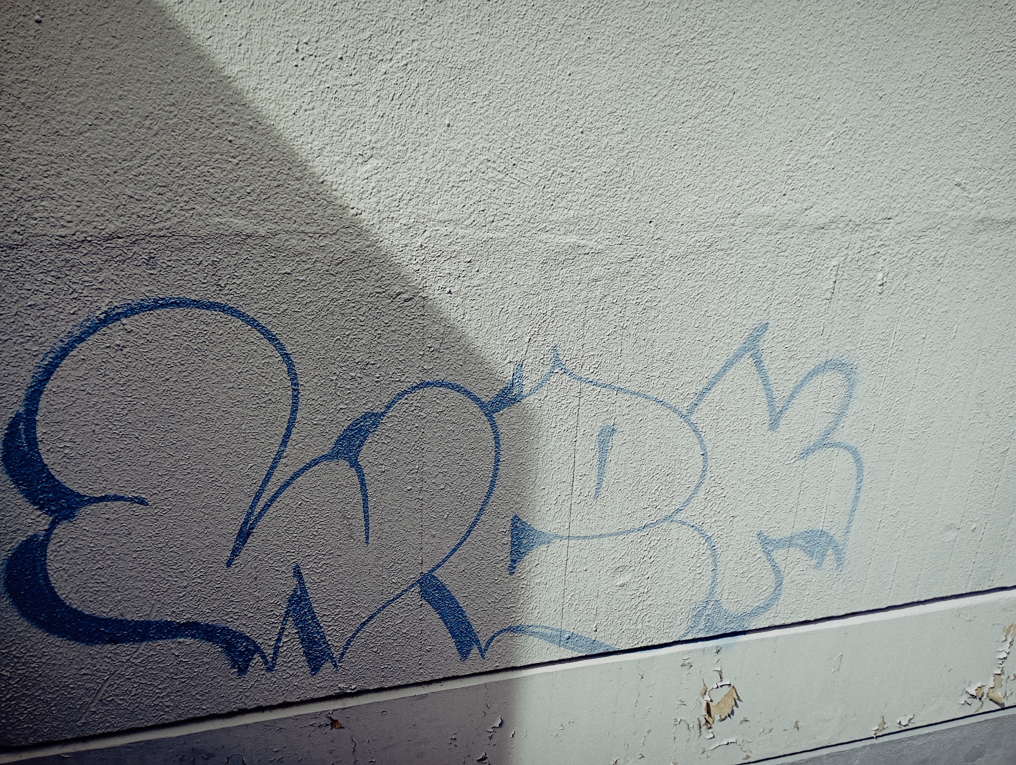 The image shows a close-up of a weathered wall with blue graffiti. The wall is textured, with a light-colored, possibly peeling layer over a concrete base. The graffiti consists of bubble letters outlined in blue paint. Part of the wall is cast in shadow, creating a contrasting light and dark area. There is damage to the base of the wall, with flaking paint exposing underlying surfaces. No people are visible in the frame. The location of this image is unknown.