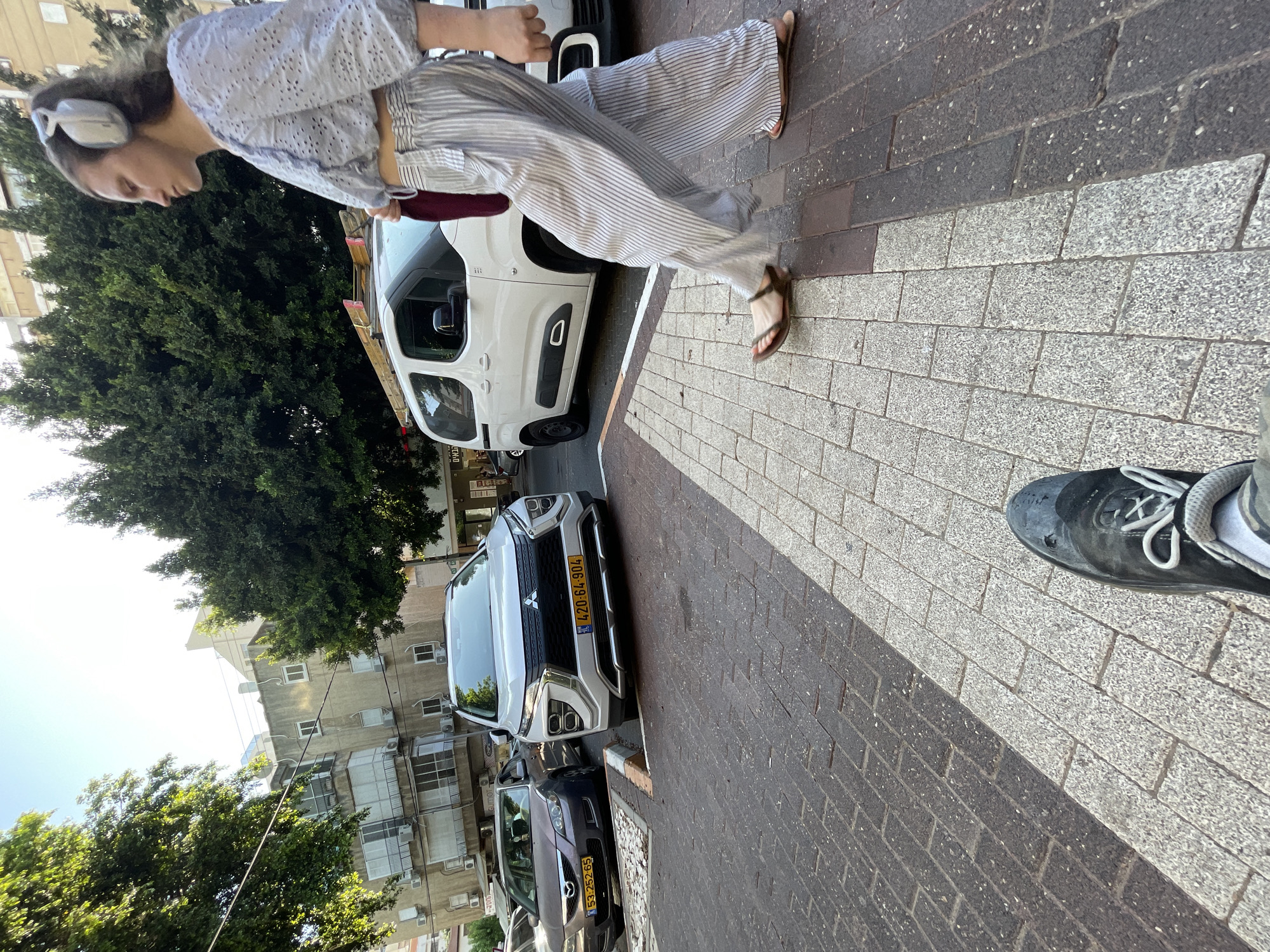 The image depicts a street scene in Kfar Saba, Israel, on a sunny day. In the foreground, a person wearing dark sneakers and grey-and-white socks is partially visible, their feet resting on a pedestrian crossing with a pattern of light and dark grey paving stones. Further down the street, a woman with brown hair, wearing noise-canceling headphones, a light blue eyelet crop top, and vertically striped wide-leg pants, is walking away from the camera towards the right. She is carrying a maroon bag in her right hand.
Behind her, two cars are parked on the street. The car on the left is a dark grey sedan with license plate 53-25265. The car on the right is a white Mitsubishi SUV with a license plate reading 420-64-904. To the right of the Mitsubishi, a white van with distinctive circular front fog lights is parked.
The background features multi-story apartment buildings with balconies and windows, interspersed with lush green trees, characteristic of a residential area in Kfar Saba. The sky is clear and bright, indicating a sunny day. The overall impression is a typical urban street environment.