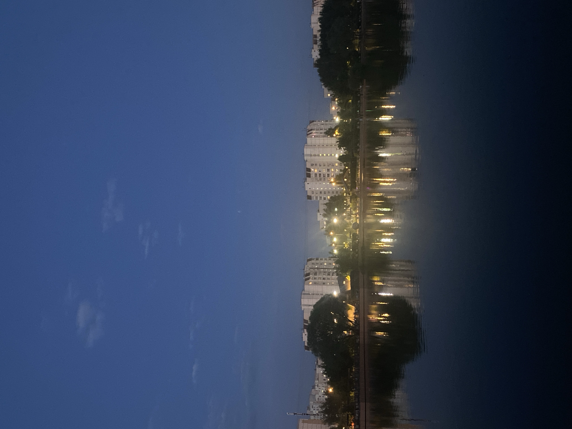 The image shows a cityscape reflected in a body of water. Buildings, trees, and lights are visible, both in their actual form and as reflections. The sky is a dark blue, suggesting it is either dusk or night. The water is calm and dark, reflecting the lights and shapes of the city. The scene suggests a tranquil, urban environment, possibly in the city of Moscow, Russia. There are no people visible in the image.