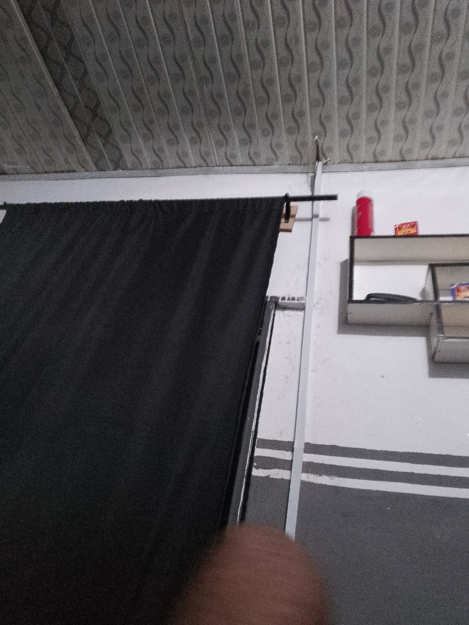 The image shows an indoor scene with a black curtain on the left and a white wall with a shelf on the right. The ceiling appears to be a corrugated material with a wavy pattern. On the shelf, there is a red cylindrical object, possibly a can or bottle, and a small red and yellow box. Below the shelf, there are two horizontal stripes on the wall, one light gray and one dark gray. A metallic pole runs vertically near the curtain. A hand is partially visible in the lower right corner of the image. The scene is dimly lit.
