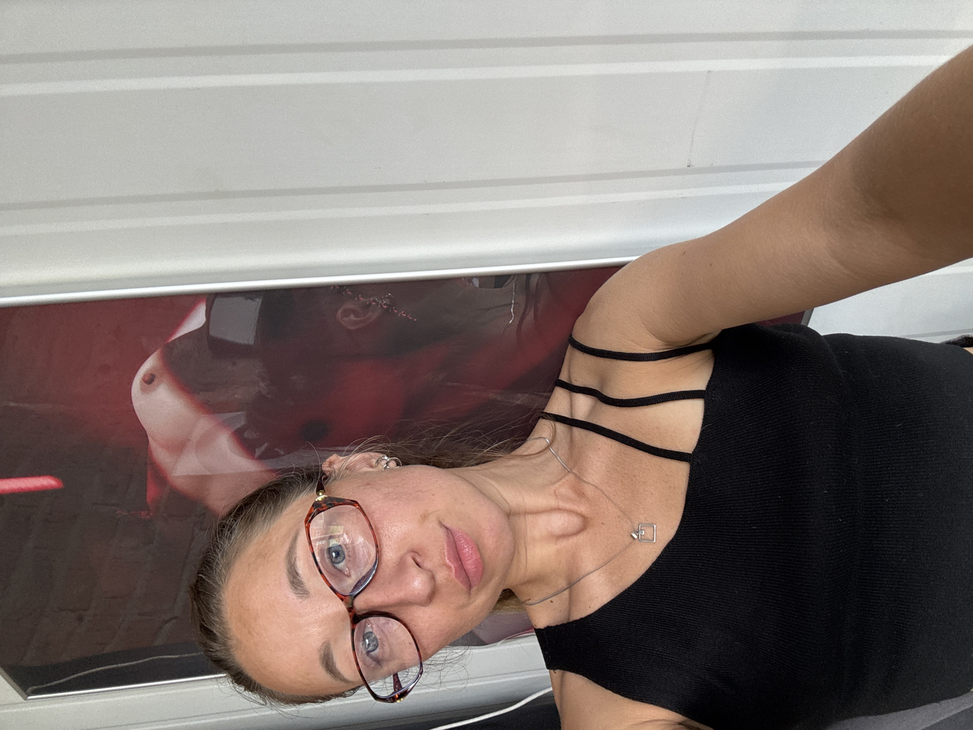 A woman with light skin, wearing glasses and a black tank top with multiple straps across the shoulders, poses indoors. She has light brown hair pulled back and is wearing a necklace with a small square pendant. Behind her is a framed picture with a red and dark background featuring a figure in an abstract manner. The picture is mounted on a white wall with horizontal grooves. The location is possibly in Russia, as the request included Chaikovsky, Russia.