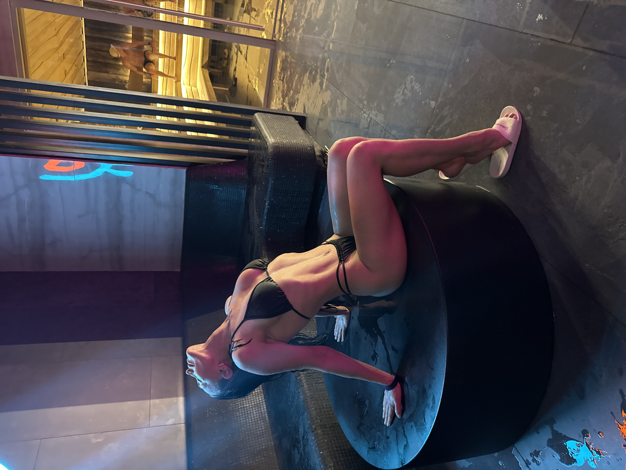 The image depicts an indoor scene, likely a spa or bathhouse. A woman is the central subject, lying on her back on a large, dark, circular platform. She is wearing a black two-piece bikini, and her body appears wet and toned. Her head is tilted back, and her arms are extended to her sides, hands resting on the wet platform. She is wearing white flip-flops, one of which clearly displays the text "Calvin Klein."
In the background, a person (partially visible from the waist down, wearing dark swimming trunks) is standing in a brightly lit, wooden-paneled area, possibly within a sauna or a shallow pool. The floor surrounding the woman is dark and appears wet, reflecting various artificial lights. A wall in the mid-ground features a bright blue neon stylized character or symbol. Further in the foreground, vibrant orange and teal splashes of color are reflected on the wet, dark tiled floor. The overall lighting is dim and atmospheric, characteristic of a contemporary spa setting. The wet surfaces and attire indicate a humid or water-based environment.