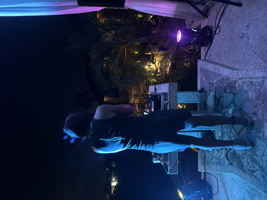 The scene depicts an outdoor nighttime event in Uluwatu, Indonesia, centered on an elevated stage where a DJ is performing.
A male individual, facing away from the viewer, stands centrally on a tiled platform, operating a DJ setup. He has long, dark hair exhibiting motion blur, wears a dark sleeveless tank top with a light vertical design on the back, dark shorts, and light-colored sneakers. His arms are raised, and his head is slightly tilted, indicating active engagement with the equipment.
The DJ setup consists of a rectangular, light-colored stone console. On the console are a DJ mixer and two media players, identifiable by their jog wheels and displays. A beverage can is visible on the left side of the console. The platform is illuminated by two stage lights: one on the left casts a prominent blue light onto the DJ and the stage, while another on the right emits purple light. A large dark speaker is partially visible on the far right, next to a flowing sheer white curtain illuminated by the purple light.
Below the elevated stage, a lush tropical environment is visible, featuring palm trees and other foliage illuminated by warm yellow ground lights. A body of water, possibly a pool or pond, reflects some of the ambient light directly in front of the stage. Further back, a group of indistinct individuals is gathered on a lower level, suggesting an audience or attendees at a social event. Buildings with illuminated interiors or exteriors are visible in the mid-ground and distant background under a dark night sky.