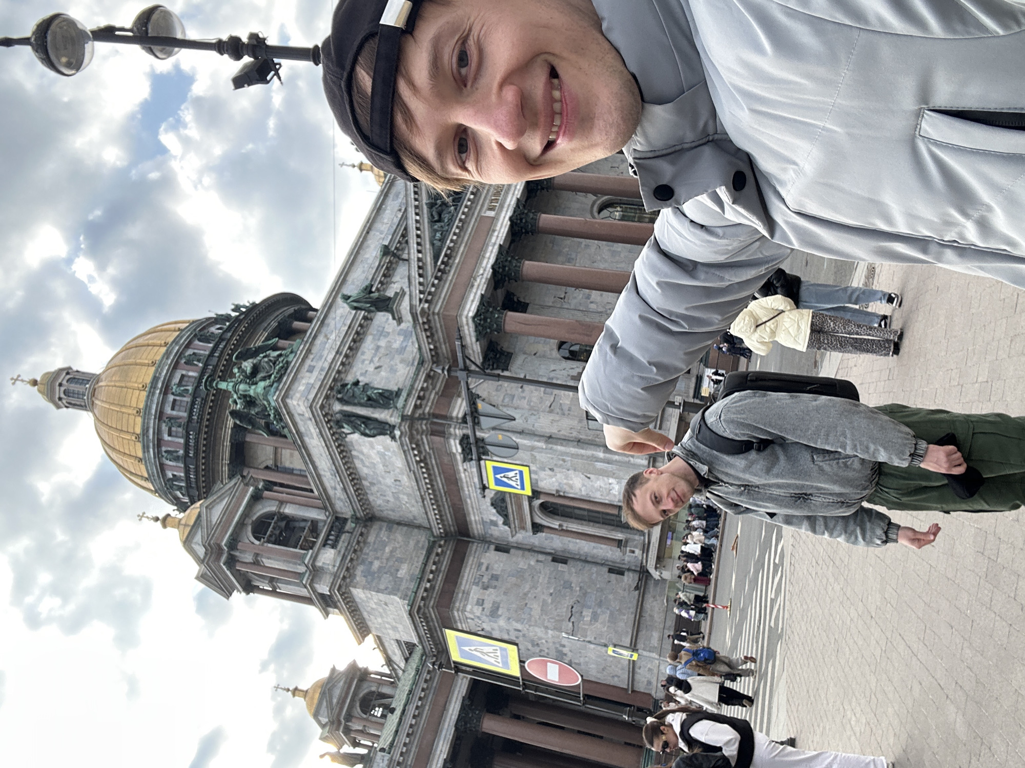 A rotated medium shot depicts two men in Saint Petersburg, Russia, with Saint Isaac's Cathedral in the background.
The first man is in the top right of the frame. He smiles at the camera and wears a black baseball cap and a light gray jacket. The second man stands at the bottom center of the image and wears a gray jacket, green pants, and a black backpack. Pedestrians walk on the sidewalk near a crosswalk in front of the cathedral. Blue and yellow crosswalk signs are affixed to the cathedral wall. The sky above the cathedral is filled with gray and white clouds. A black lamppost is in the upper left corner.