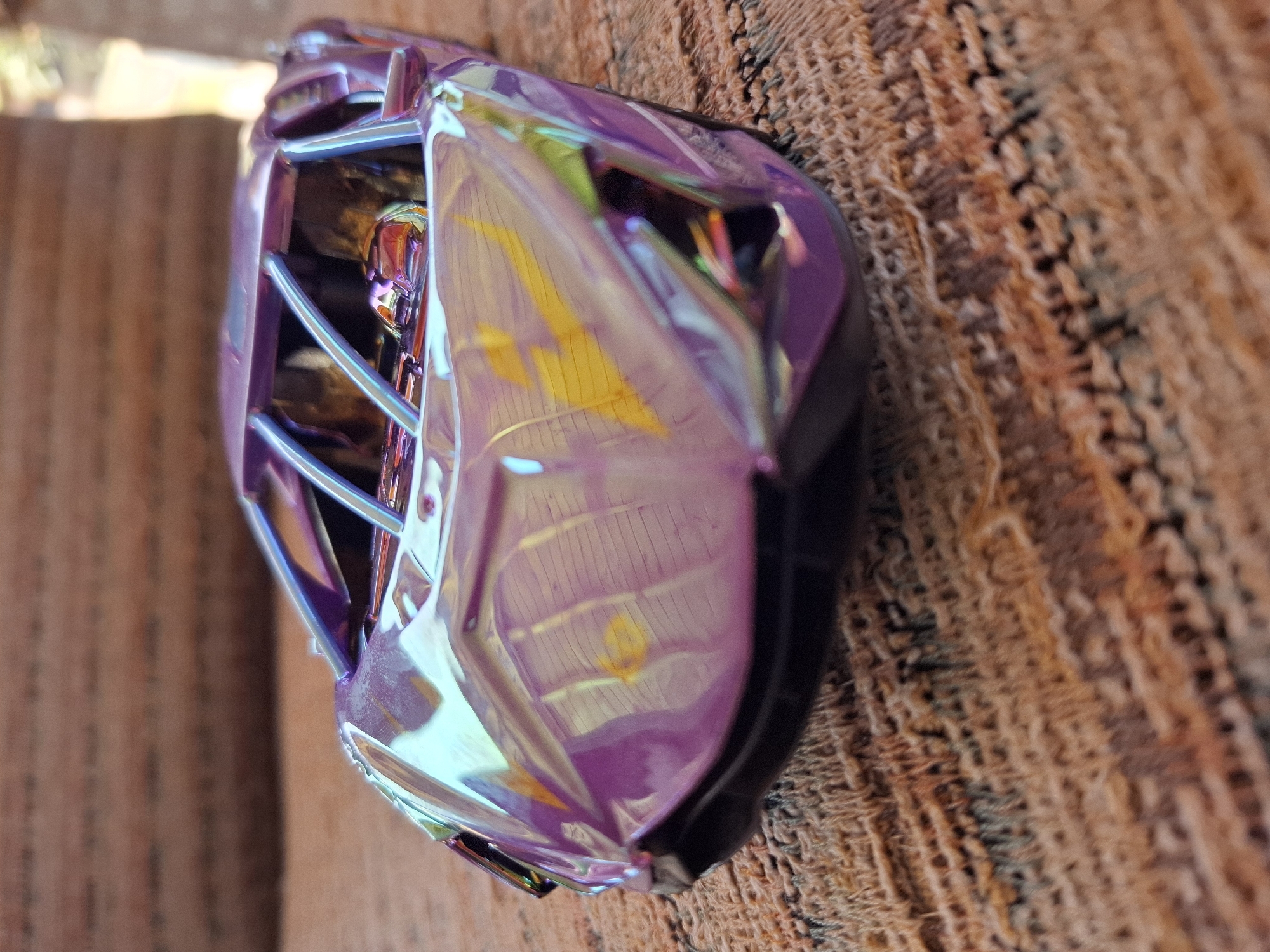 A close-up, low-angle, slightly blurry shot captures a small, iridescent purple toy car parked on a textured, brown woven surface, possibly a rug or upholstered furniture. The car's metallic finish reflects light in streaks of yellow, green, and blue. The car appears to be a sports car model, with a low profile and visible interior roll cage structure. The background is out of focus, showing muted colors that suggest an indoor environment with some natural light filtering in from the right. There are no people or animals visible in the image. The specific location in Galabovo, Bulgaria, is not discernible from the visual information provided in the image.