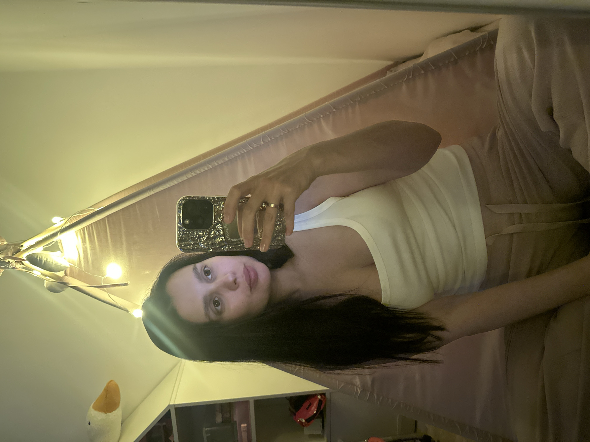 The image shows a woman holding a smartphone in a selfie pose, indoors. She is fair-skinned with dark hair and is wearing a white tank top and beige pants. Her phone has a patterned case and she is wearing a gold ring on her finger. Behind her is what appears to be a children's play tent with string lights and a plush goose decoration. There is a shelf with various items on it visible in the lower part of the frame. There is no information to identify the location as Chaikovsky, Russia.