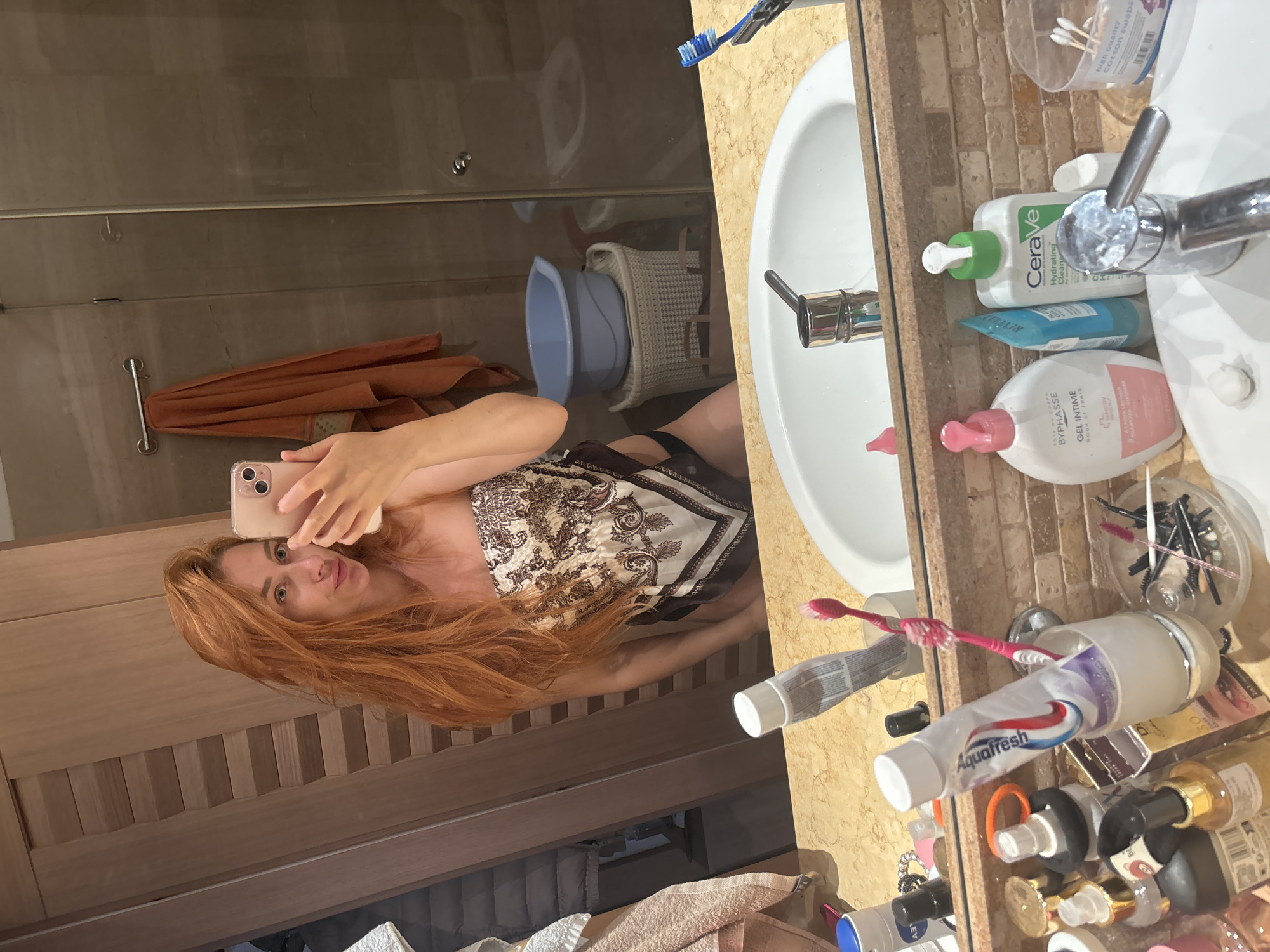 A young woman with long, auburn hair is taking a selfie in a bathroom mirror. She is wearing a strapless top with a paisley print and dark bikini bottoms. Her left arm is raised, holding a pink smartphone. The bathroom features wooden cabinetry, a marble countertop with a white sink, and a large mirror reflecting the scene. Various toiletries, including toothpaste, toothbrushes, and bottles of skincare products, are arranged on the countertop. In the background, a towel hangs on a rack, and a blue plastic basin sits atop a woven basket. The overall impression is a personal, candid moment captured in a domestic setting.