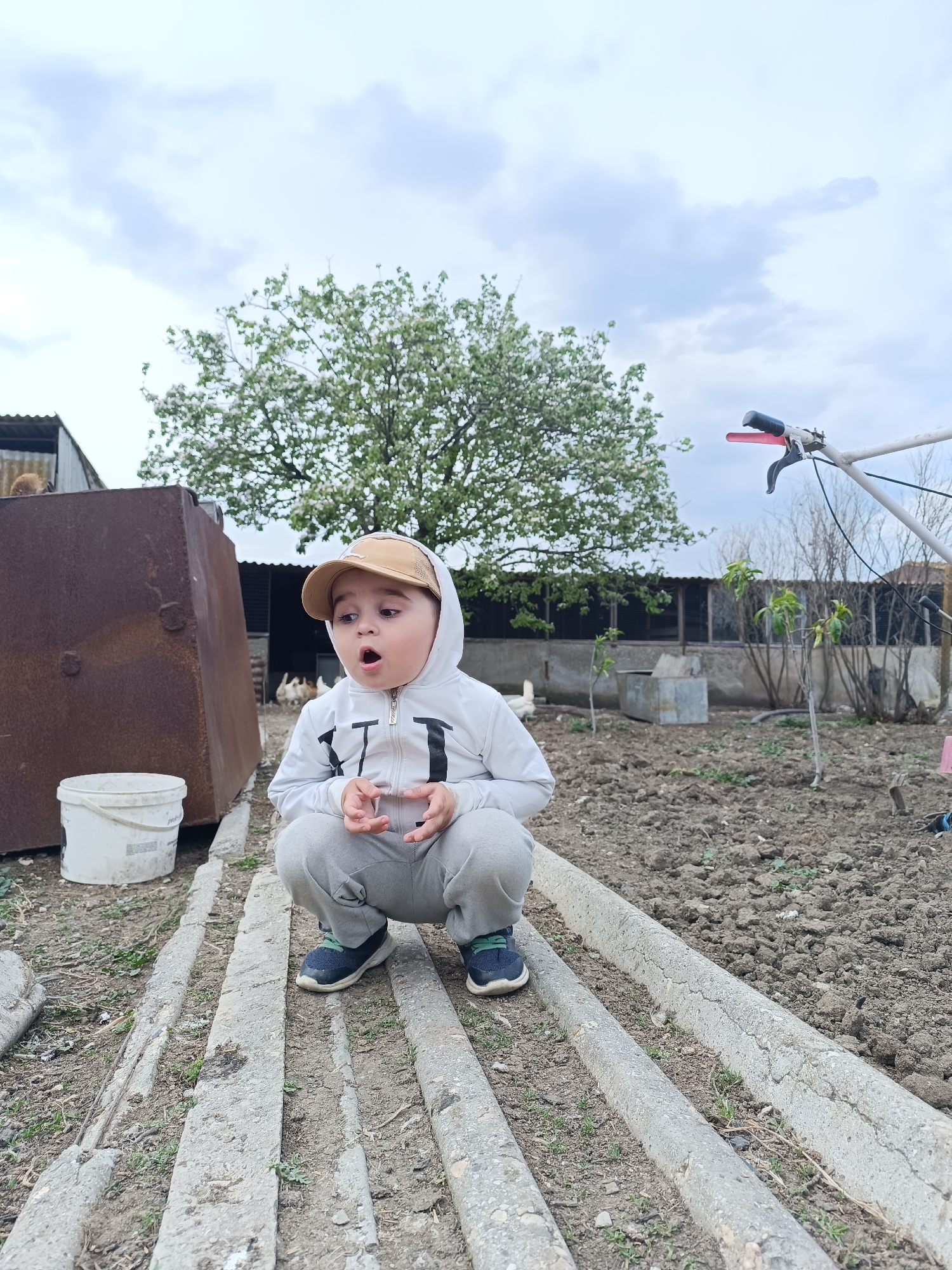 A child squats in an outdoor environment. The child wears a white hooded sweatshirt, gray sweatpants, and a tan baseball cap. The child's mouth is open in an expression of surprise. They are positioned on several parallel concrete blocks or beams. Behind the child is a garden or plot of land, a large tree with white flowers, and what appears to be a chicken coop structure with several chickens inside. To the left is a rusted metal container next to a white bucket. In the background, there is agricultural equipment. The photo was taken in Зыбинское сельское поселение, Ukraine.