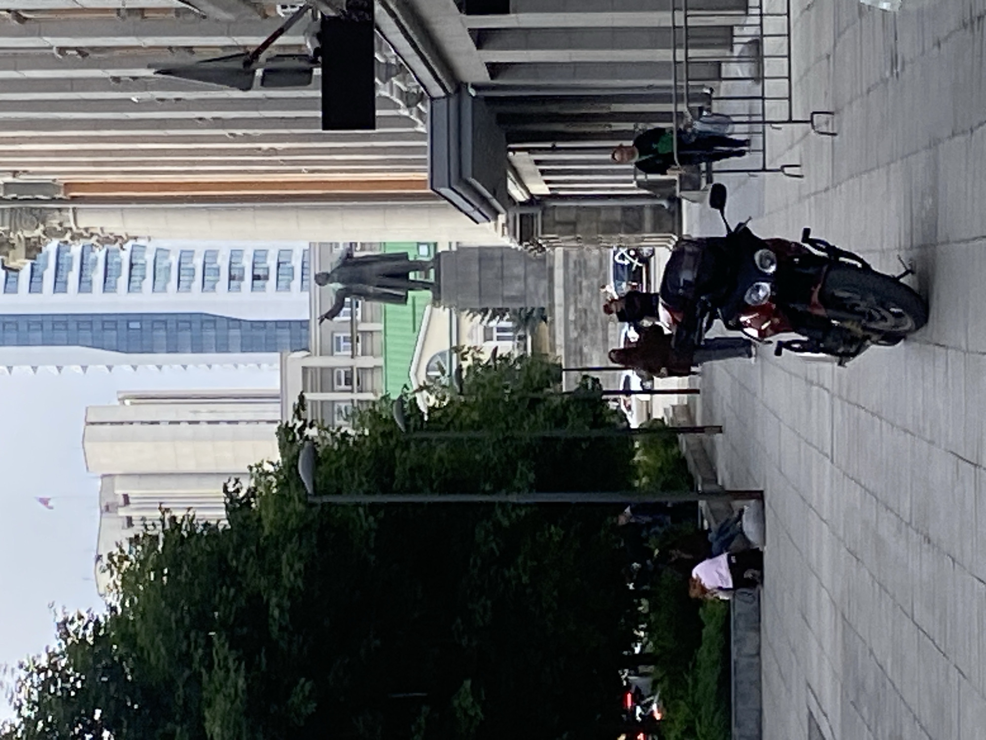 The image captures an urban street scene in Yekaterinburg, Russia, during what appears to be a bright, overcast day. The perspective is from a slightly elevated vantage point, looking across a wide, paved pedestrian area or square.
A central feature in the mid-ground is a dark-colored statue of a man, characteristic of Vladimir Lenin, standing on a pedestal with his right arm extended, pointing ahead. Behind the monument, the city's architecture showcases a blend of styles, from older buildings with ornate details and green rooftops to modern, multi-story high-rises with numerous windows, extending into the background.
On the right side of the paved area, a red and black motorcycle is parked. Several people are visible in the vicinity of the motorcycle; one individual appears to be engaging with it, while others are standing nearby. Further to the right, behind a black metal barrier, a person in a green shirt stands. Towards the foreground on the right, another individual wearing a light-colored top is seated on a low stone ledge or wall. Numerous other pedestrians are scattered throughout the scene, suggesting a typical day in a busy city center.
Green foliage from trees lines parts of the street, contrasting with the concrete and buildings. In the far background, a string of colorful flags or banners can be faintly seen against the bright sky. The lighting indicates daytime, likely late morning or afternoon, with no harsh shadows, consistent with an overcast sky. No discernible text is visible in the image.
