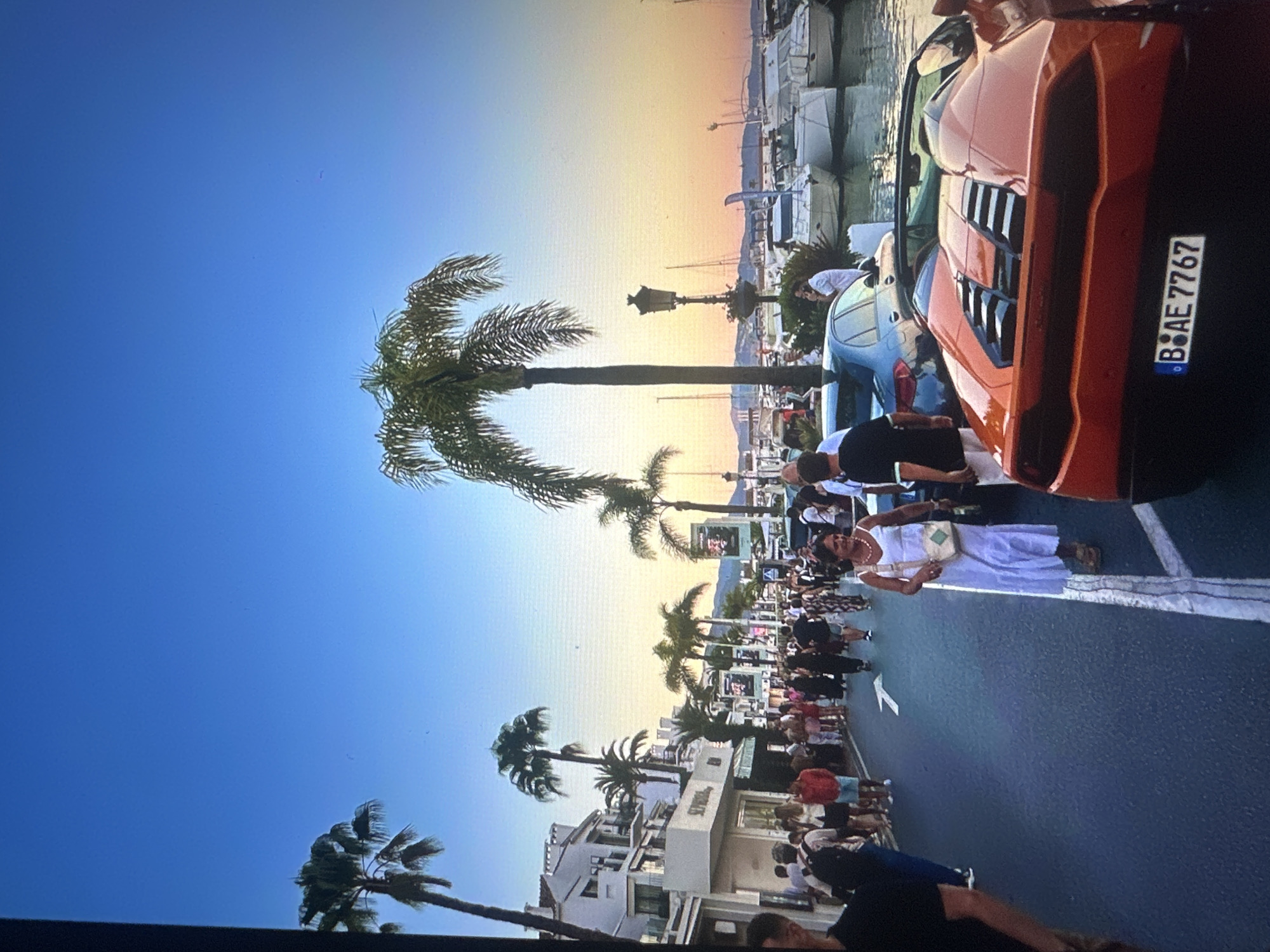 The image depicts a street scene in a coastal city at dusk. The sky transitions from a clear blue to a warm orange and yellow gradient near the horizon, suggesting sunset. Palm trees are scattered along the street, framing a view of a harbor filled with numerous boats and yachts.
On the street, a diverse crowd of people are walking, some individually and others in groups. They are dressed in casual summer attire. A woman in a white dress is walking towards the viewer, carrying a crossbody bag. To her left, a man in a black t-shirt and shorts stands near a light blue car. In the foreground, the rear of a vibrant orange sports car is visible, with its license plate reading "B BAE 7767".
The buildings lining the street appear to be multi-story structures with white facades and balconies, indicative of a Mediterranean or similar climate. Some shops are visible on the ground floor. The overall atmosphere suggests a lively, popular destination, possibly a promenade or waterfront area in a resort town. The location provided is Tallinn, Estonia, which is not typically associated with palm trees and this kind of harbor scene, suggesting a potential misidentification of the location or a very specific microclimate.