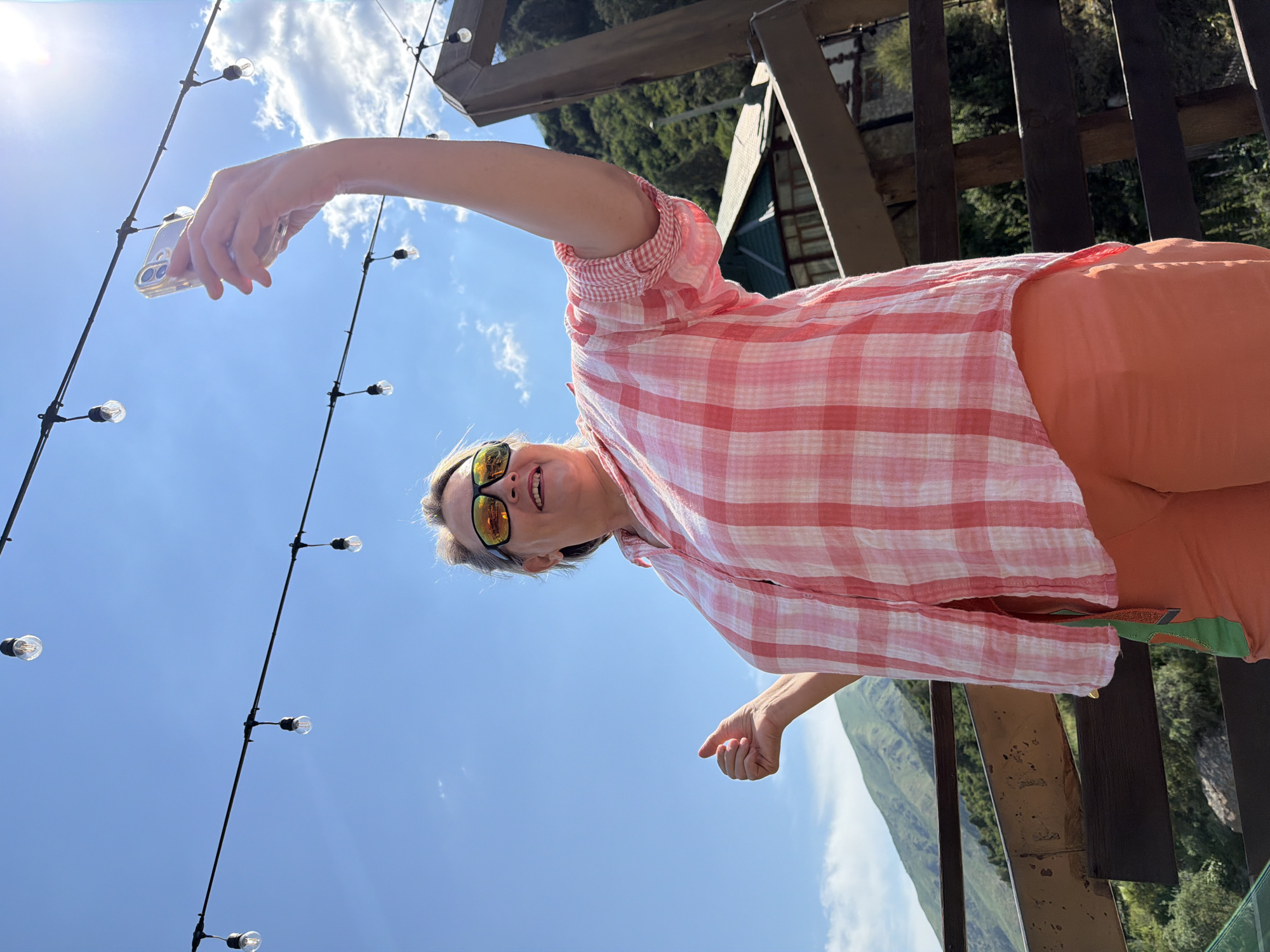 A woman is taking a selfie with a cell phone in her right hand. She is wearing sunglasses, a pink and white checkered shirt, and orange shorts. She is standing on a wooden balcony with a view of mountains and trees. There are string lights overhead.